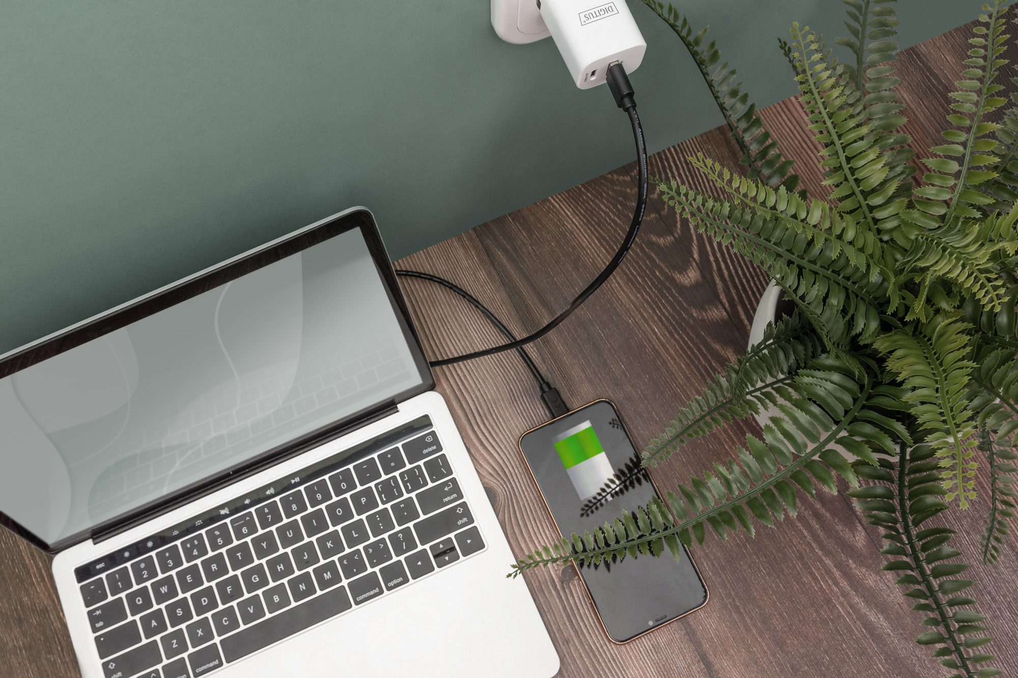 Laptop die oplaadt met een telefoon en een plant op een houten bureau. De telefoon toont de laadstatus.