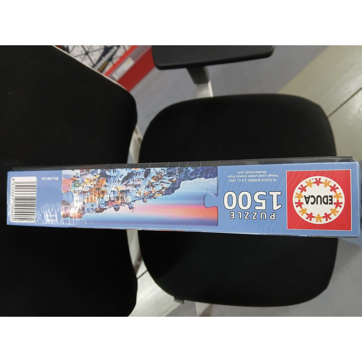 Pudełko z puzzlami, przedstawiające scenę nadmorską z niebieskimi budynkami, zachodem słońca i czarnym krzesłem.