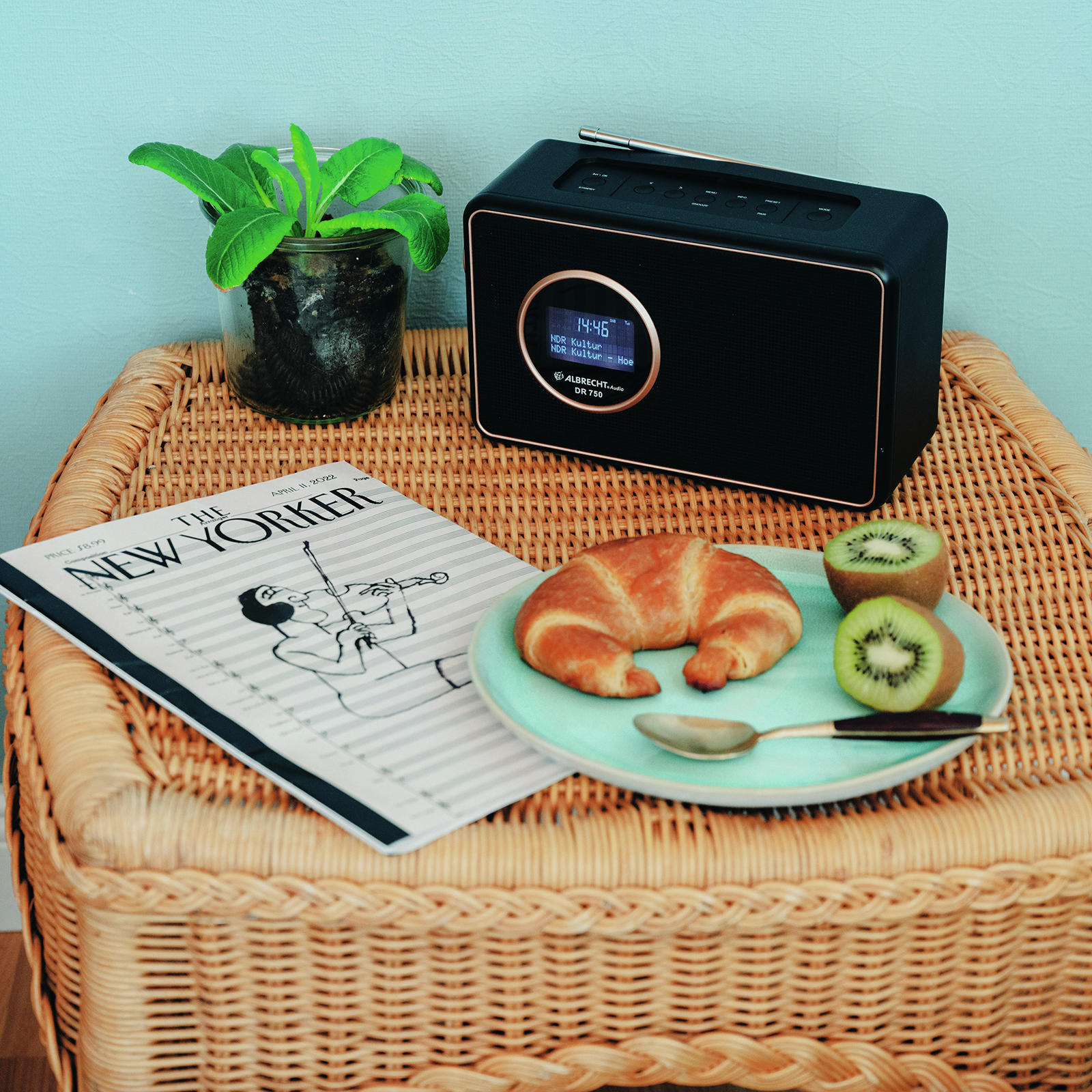 Une radio noire avec affichage numérique, un journal, un croissant, une assiette avec kiwi et une plante.