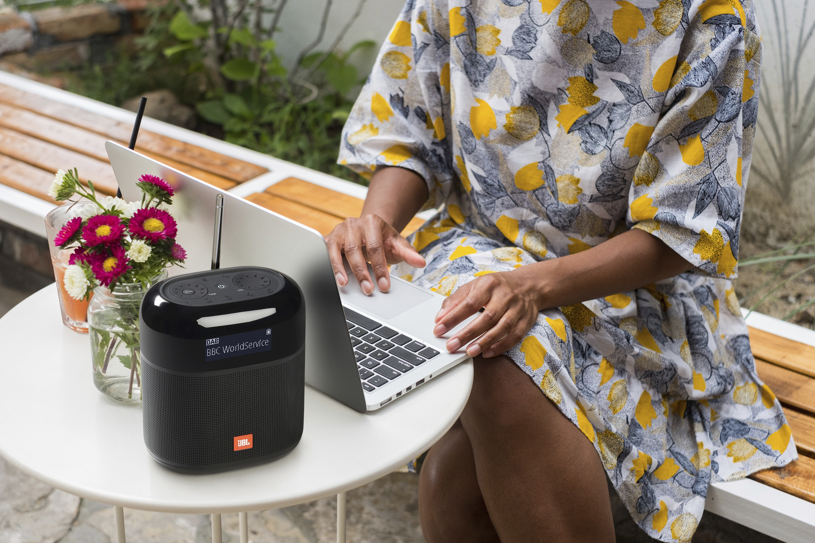 Kobieta pracuje na laptopie z głośnikiem obok, na zewnątrz. Kwiaty i napoje w pobliżu.