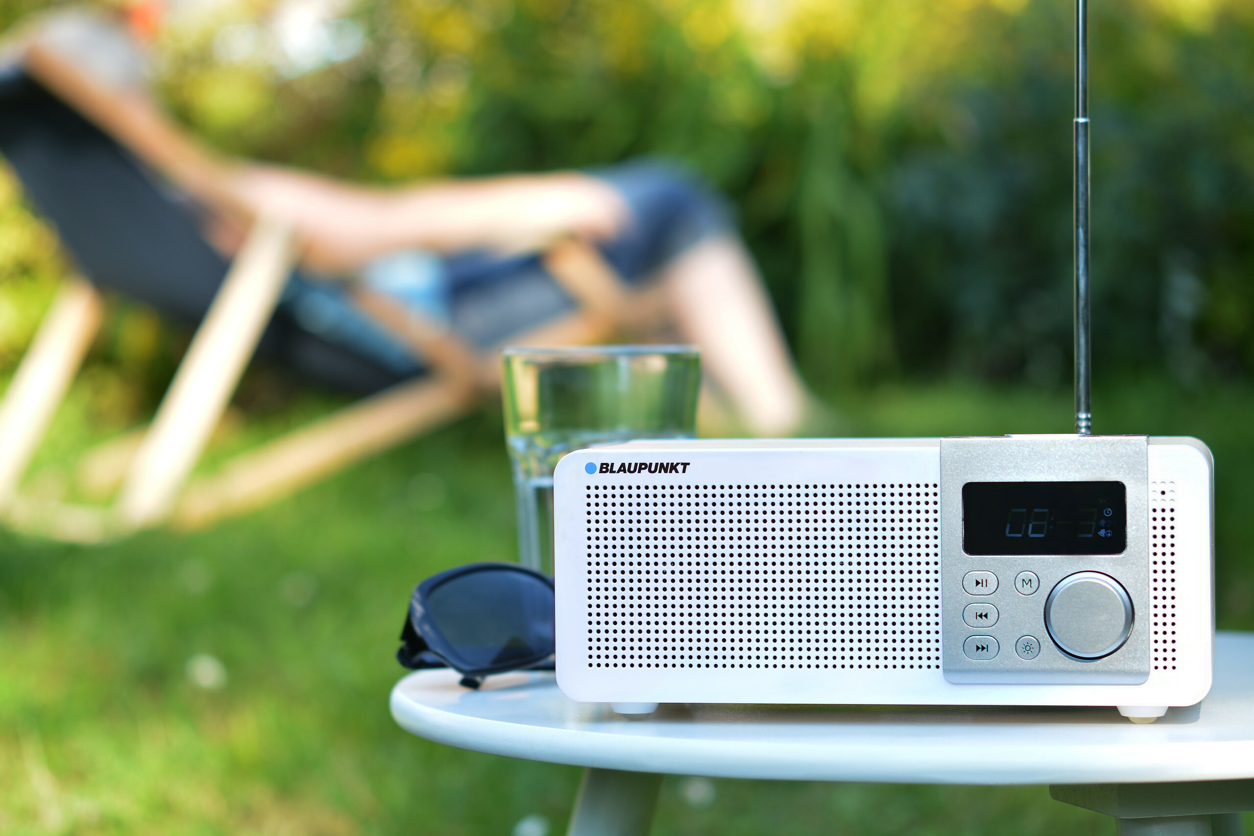 Una radio bianca con display è su un tavolo all'aperto vicino a un bicchiere e occhiali da sole.