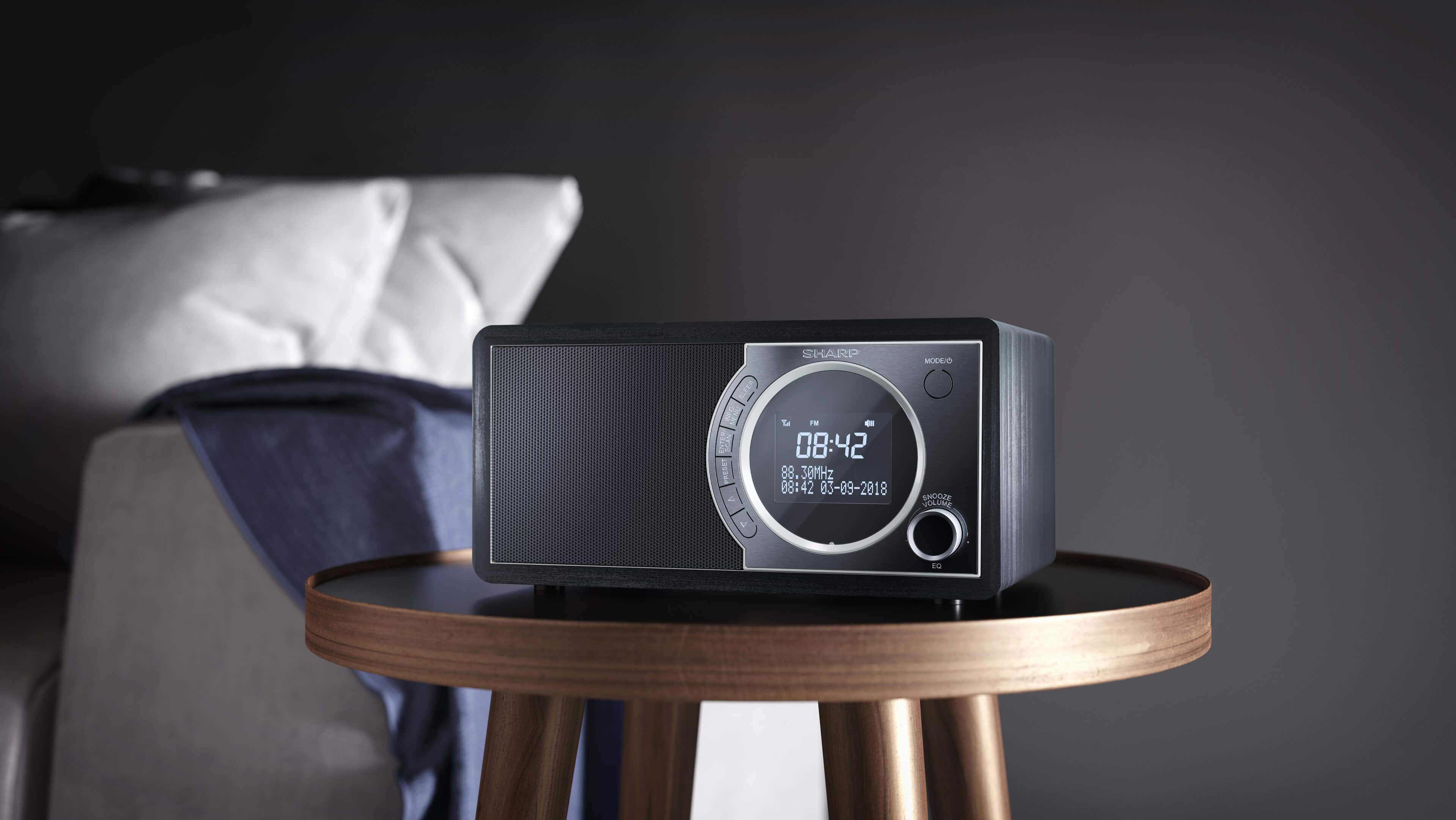 Una radio negra y plateada sobre una mesa de madera. Una cama y almohadas en el fondo borroso.