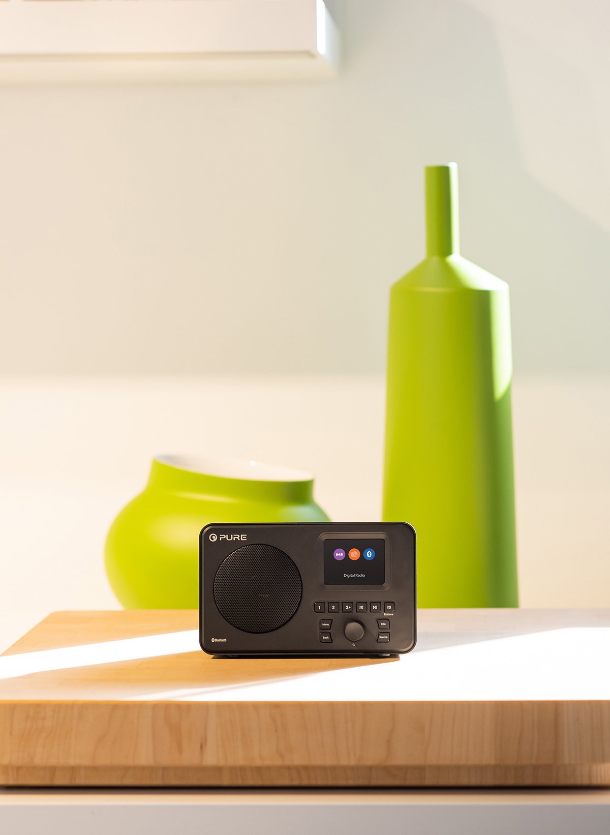 Ein schwarzes Radio auf einer Holzoberfläche, mit einer grünen Vase und Flasche im Hintergrund.
