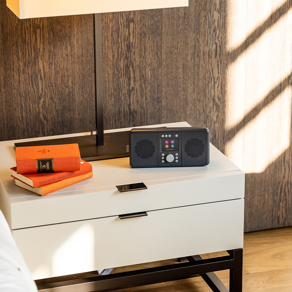 Szafka nocna z książkami i radiem, oświetlona lampą, na tle drewnianej ściany.