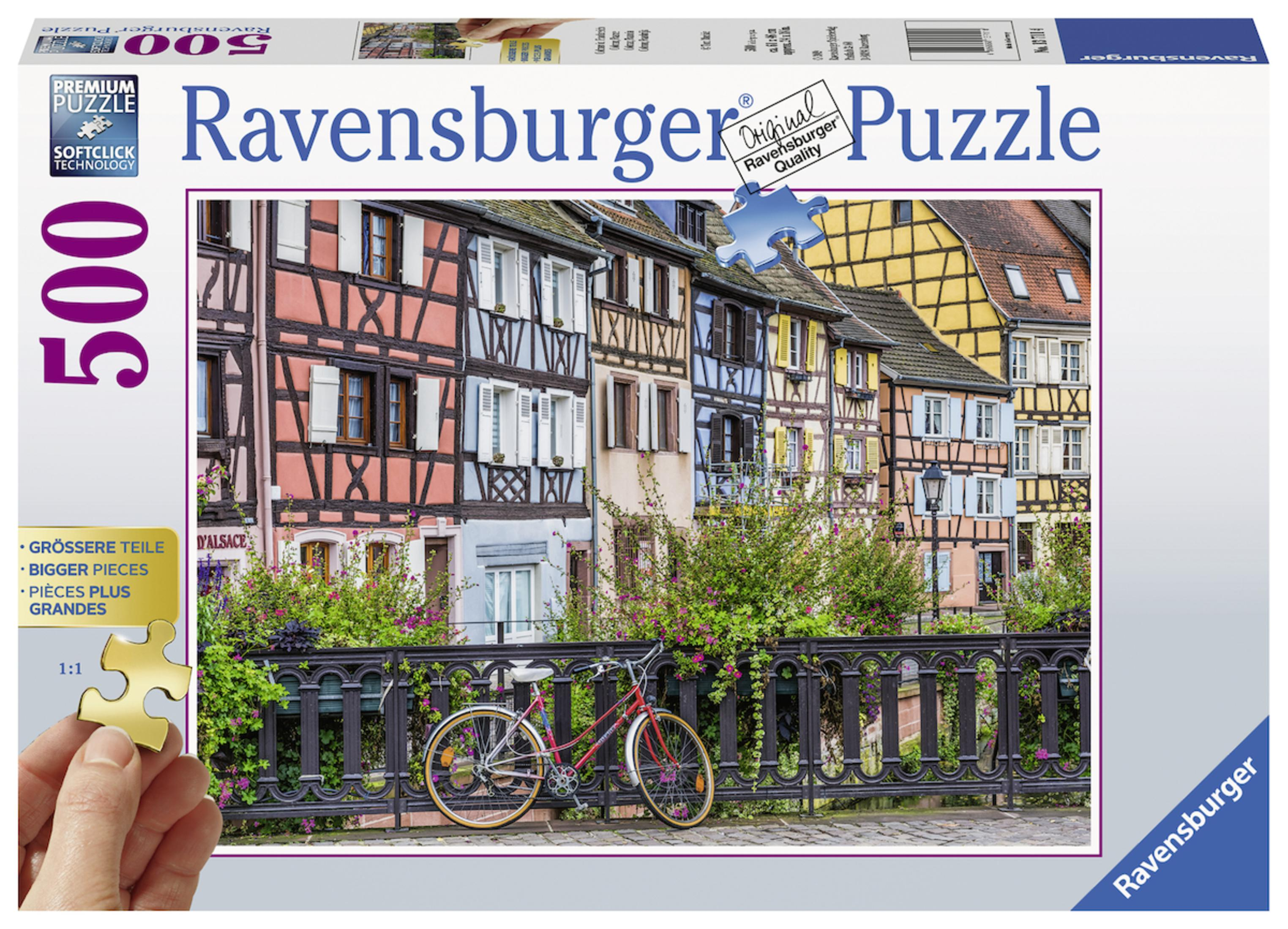Puzzelschachtel mit Bild von bunten Häusern und Fahrrad auf Brücke. Hand hält ein Puzzleteil.