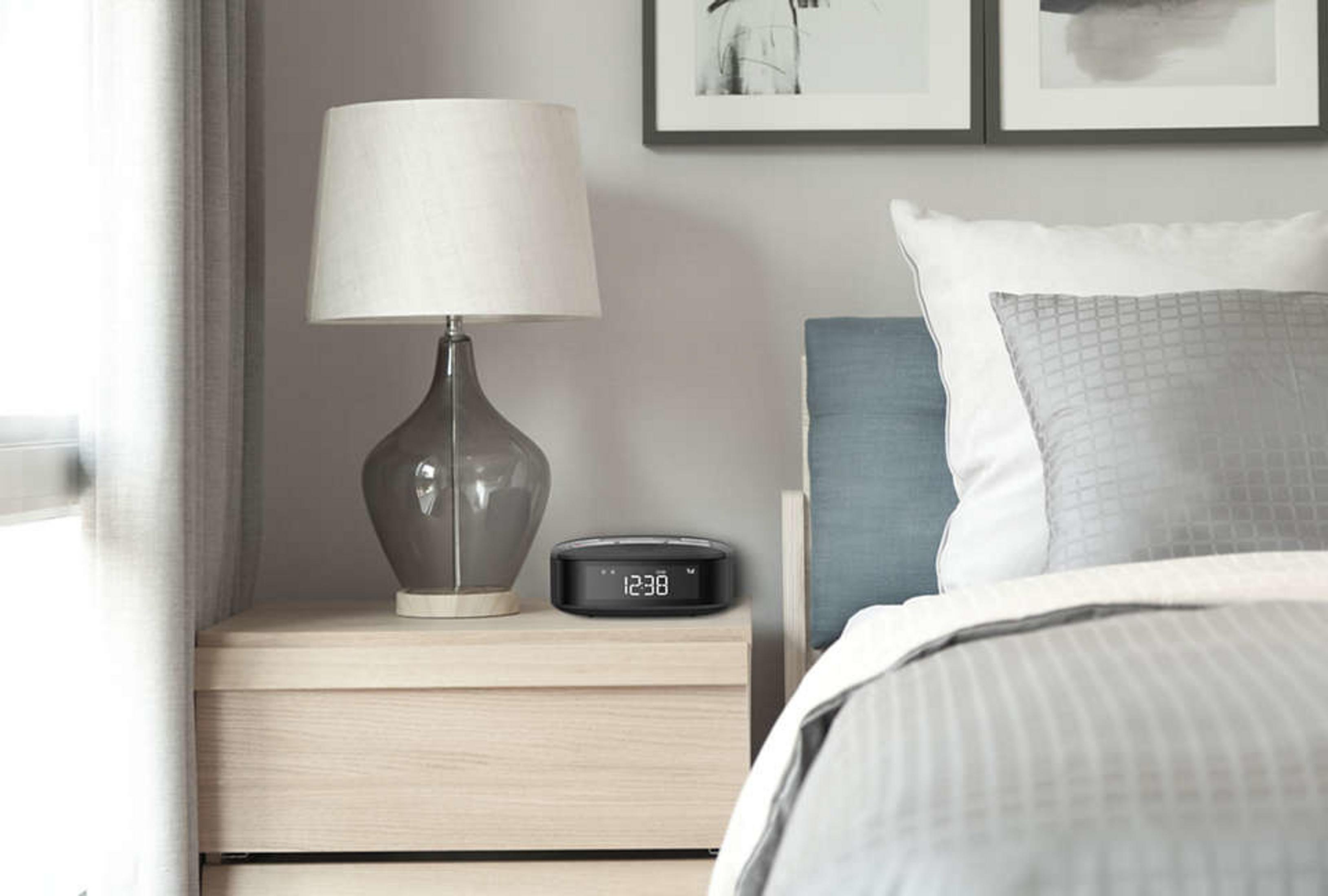 Une table de chevet dans la chambre avec un réveil Philips et une lampe. L'horloge affiche l'heure, et un lit est en arrière-plan.