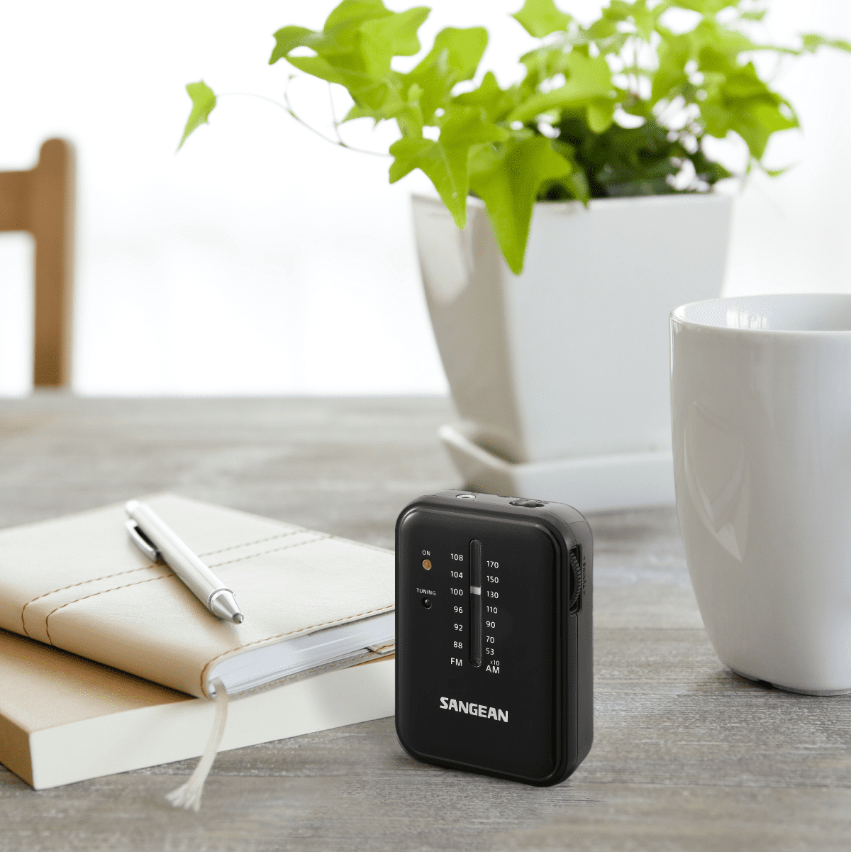 Małe czarne radio Sangean na drewnianym stole. Na stole znajdują się również notatnik, długopis i roślina.
