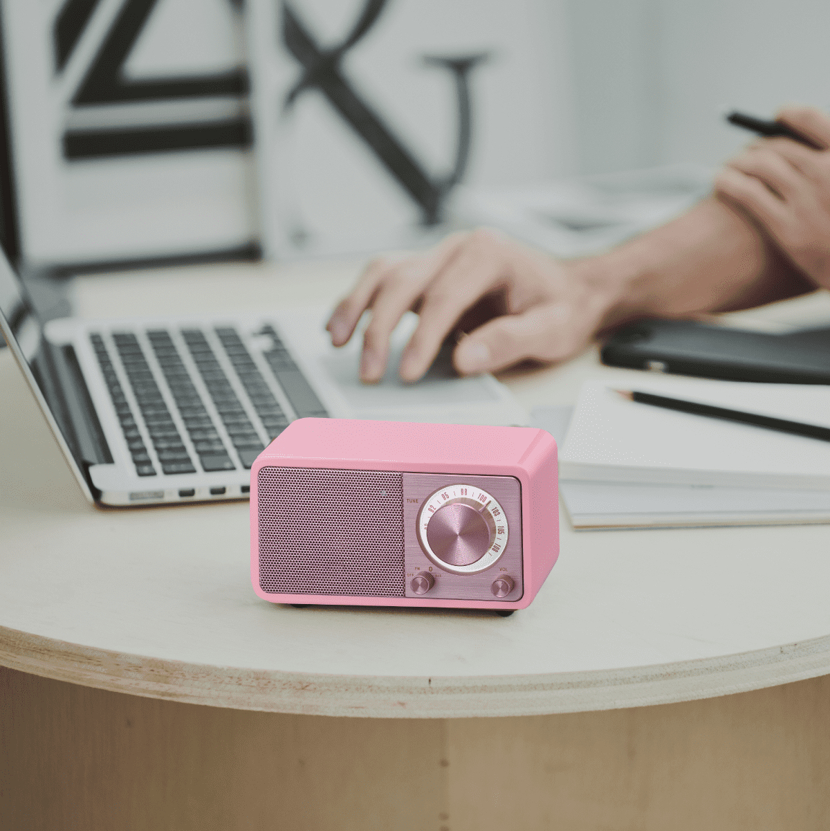 Różowe radio w stylu retro na drewnianym biurku. Osoba używa laptopa w tle. Radio jest różowe.