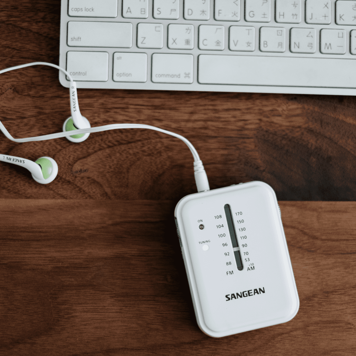 Białe radio Sangean, słuchawki i klawiatura na drewnianym biurku.