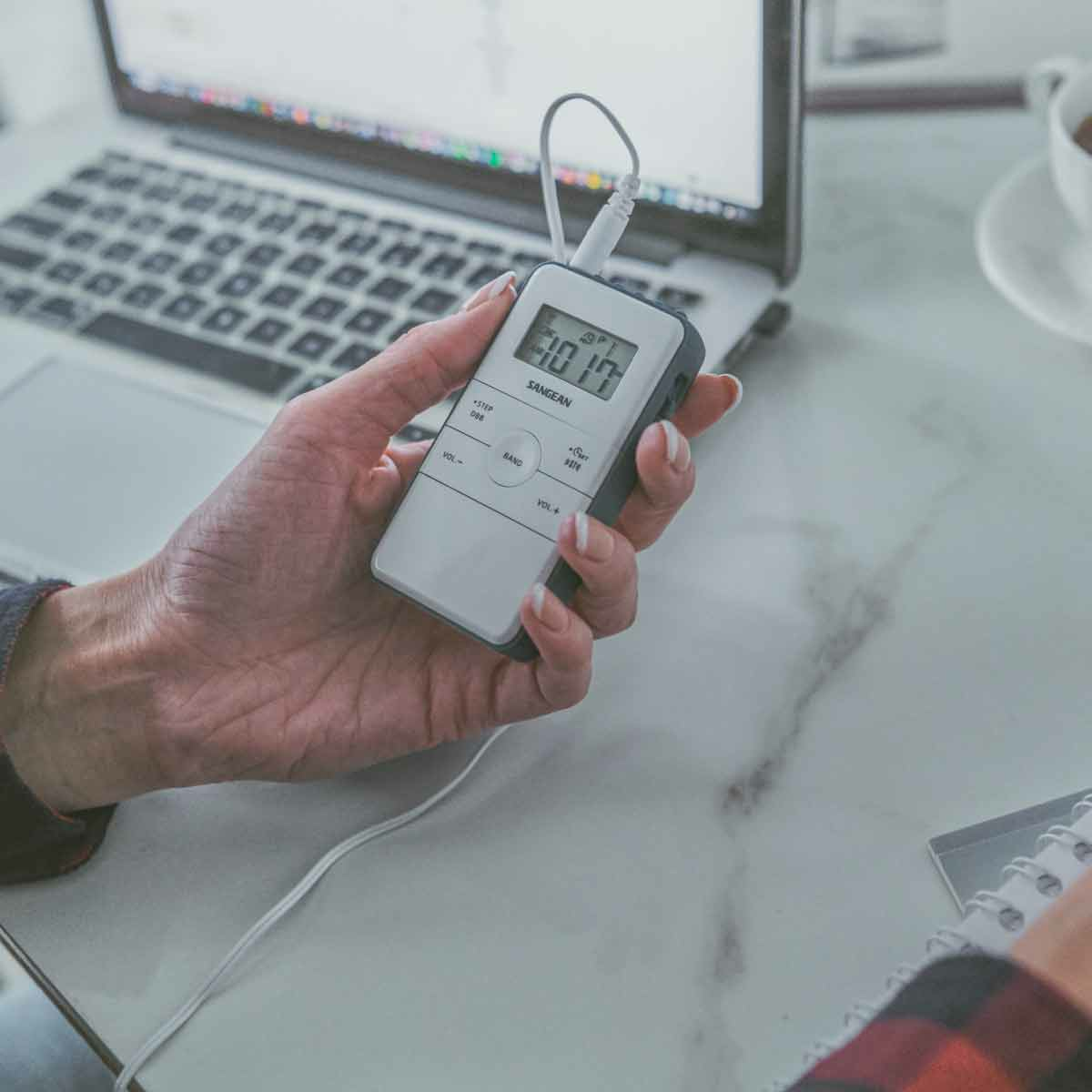 Dłoń osoby trzymającej białe urządzenie z ekranem, w pobliżu laptopa.