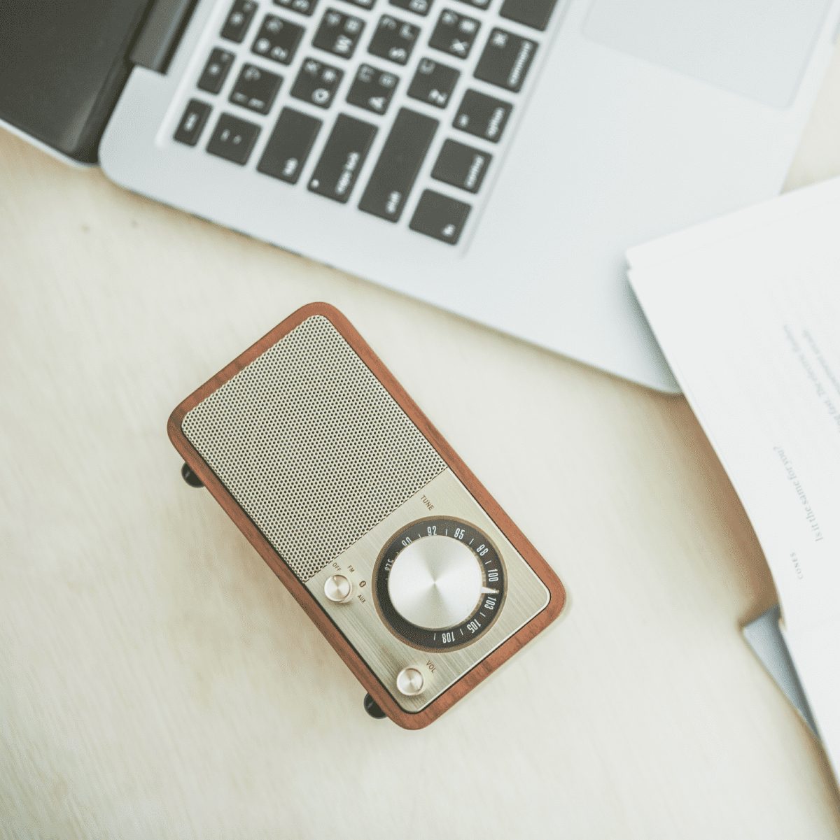Małe brązowo-srebrne radio z laptopem, papierami i długopisem na jasnym drewnianym stole. Ujęcie z góry.