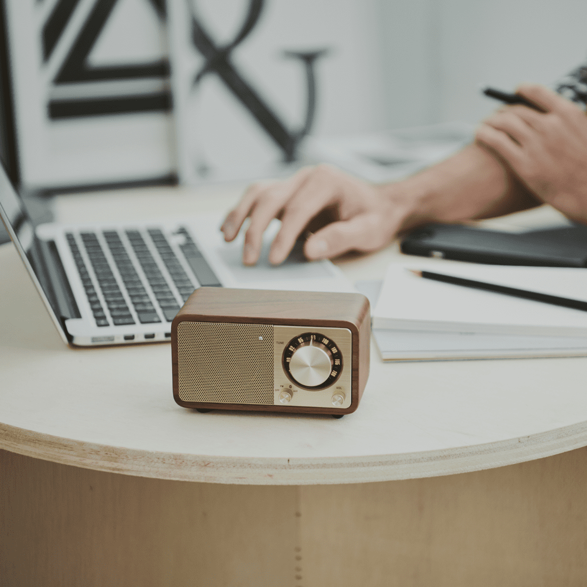 Drewniane radio na stole, w tle ręce osoby i laptop.