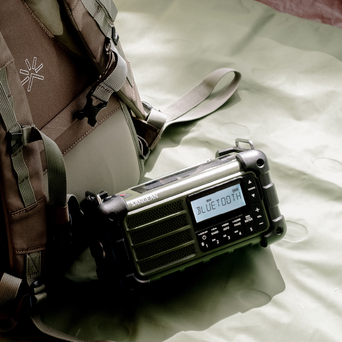 Una robusta radio Sangean Bluetooth è su una superficie verde accanto a uno zaino. La radio mostra 'BLUETOOTH'.