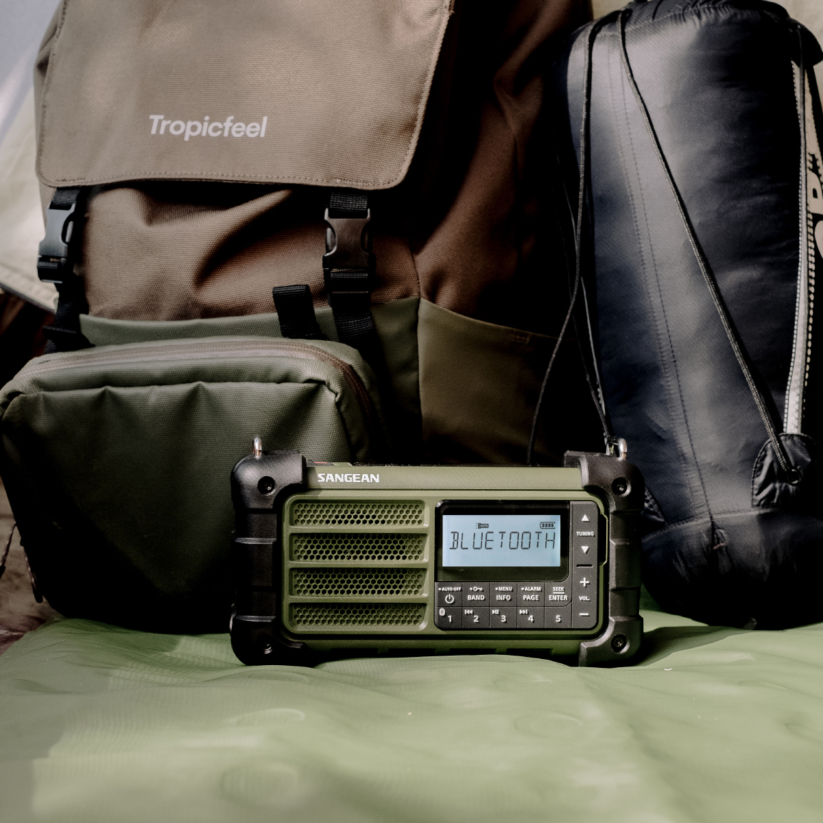 Una radio SANGEAN verde mostra 'BLUETOOTH'. Dietro, uno zaino, una borsa e un tappetino verde.