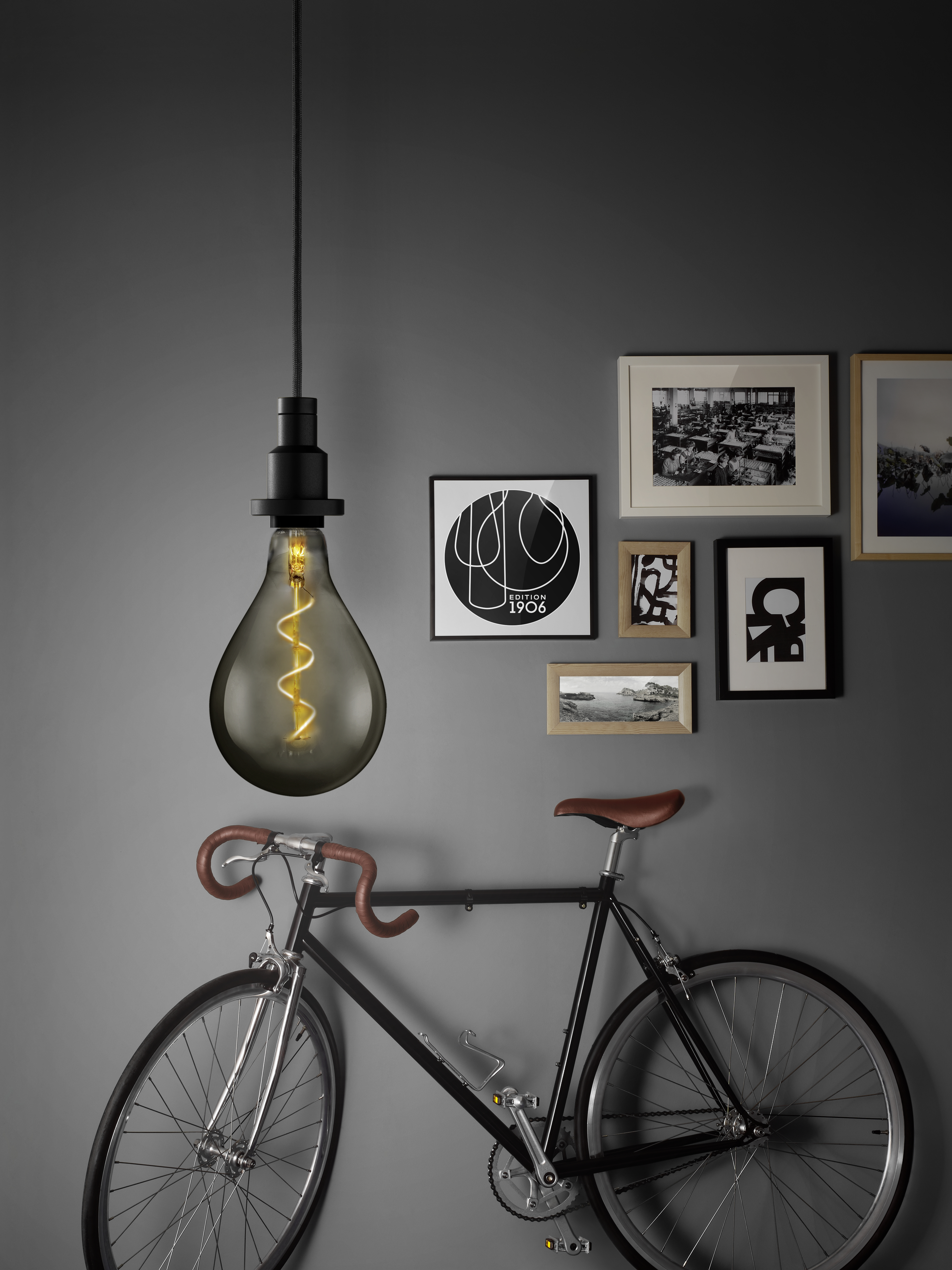 Una bicicletta nera con una lampadina sospesa e cornici su un muro grigio.