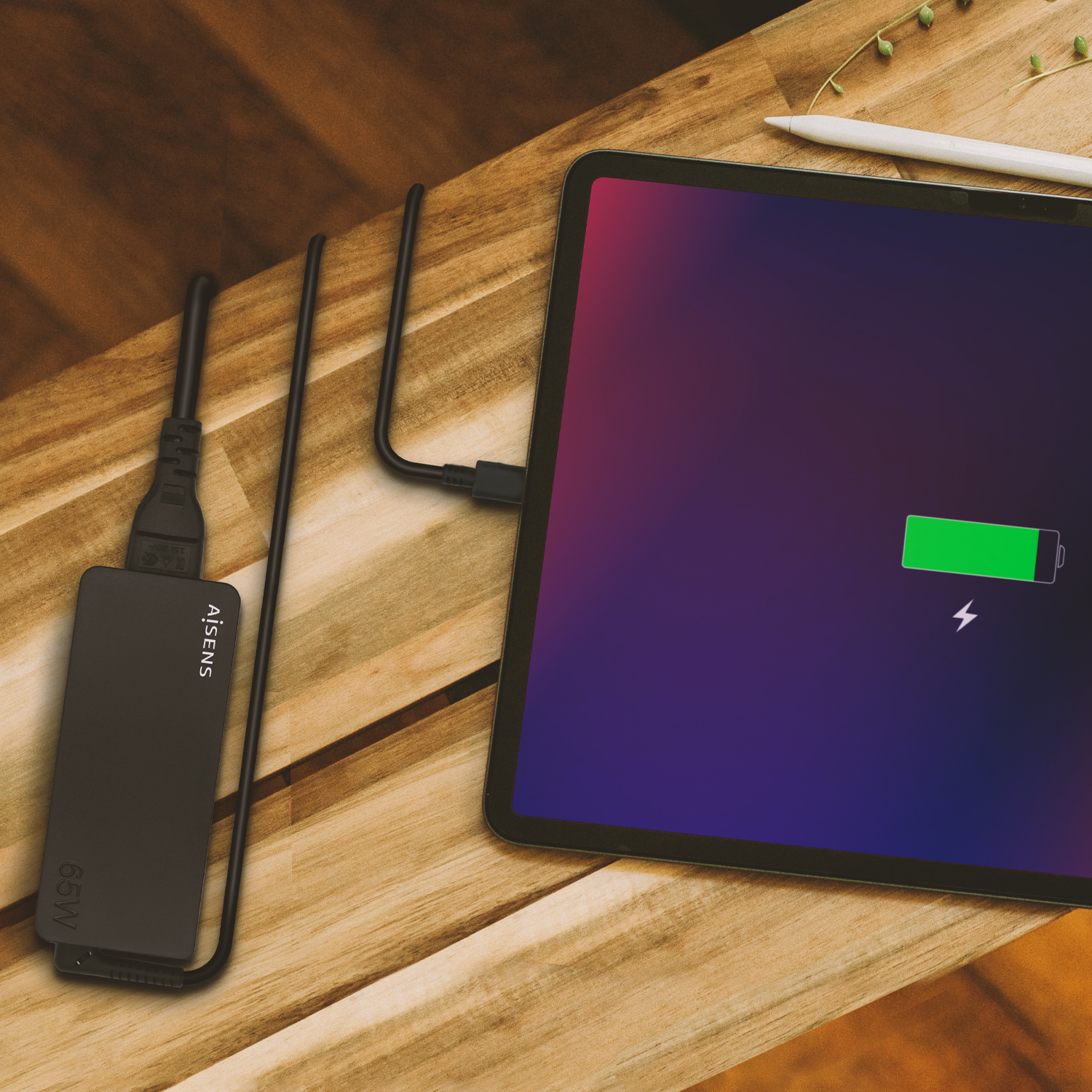 Cargador negro conectado a una tableta, cargando con un icono de batería verde en una mesa de madera.