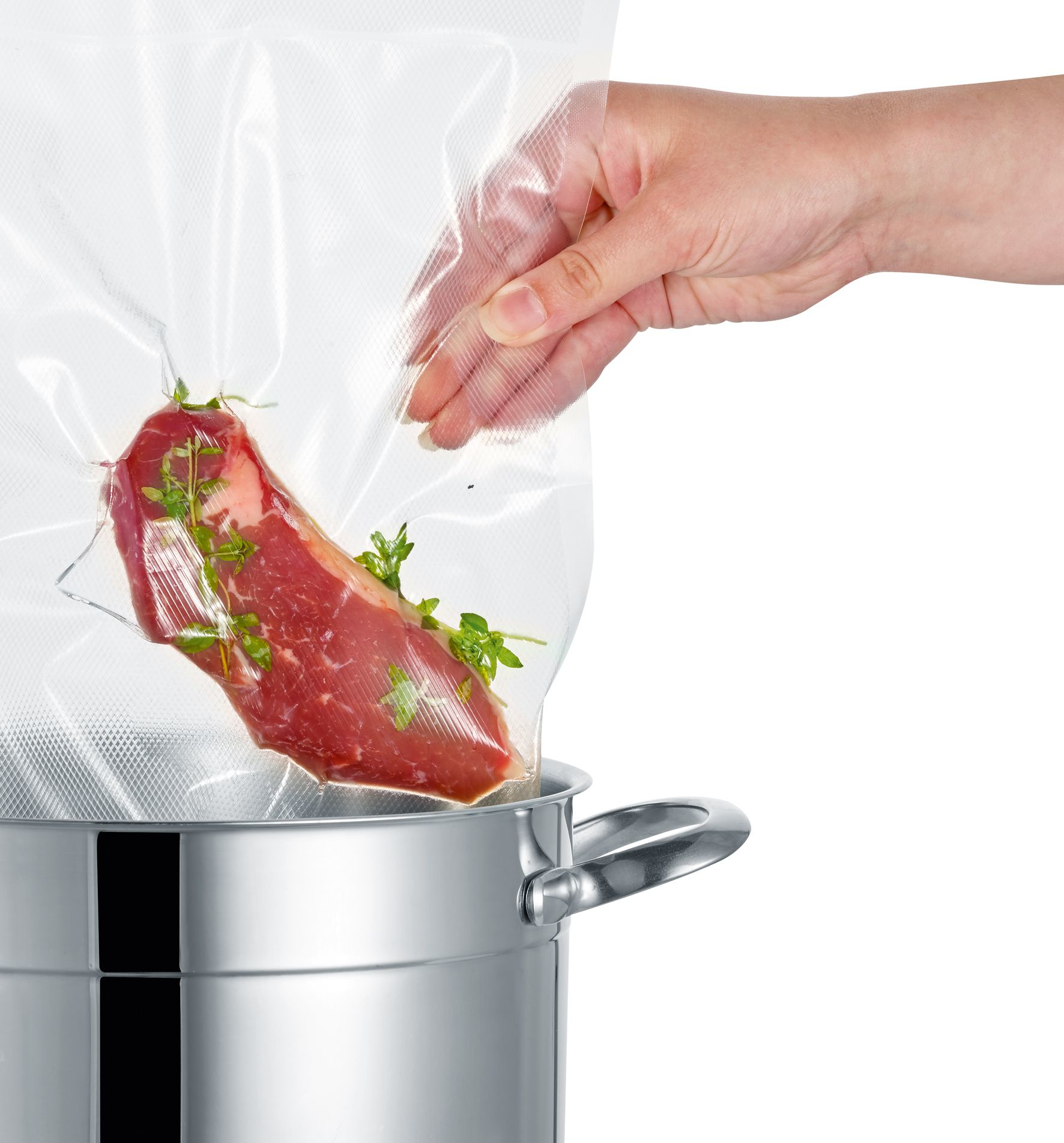 Eine Hand legt ein Stück rohes Fleisch mit Kräutern in einen Vakuumbeutel über einen Edelstahlkochtopf.