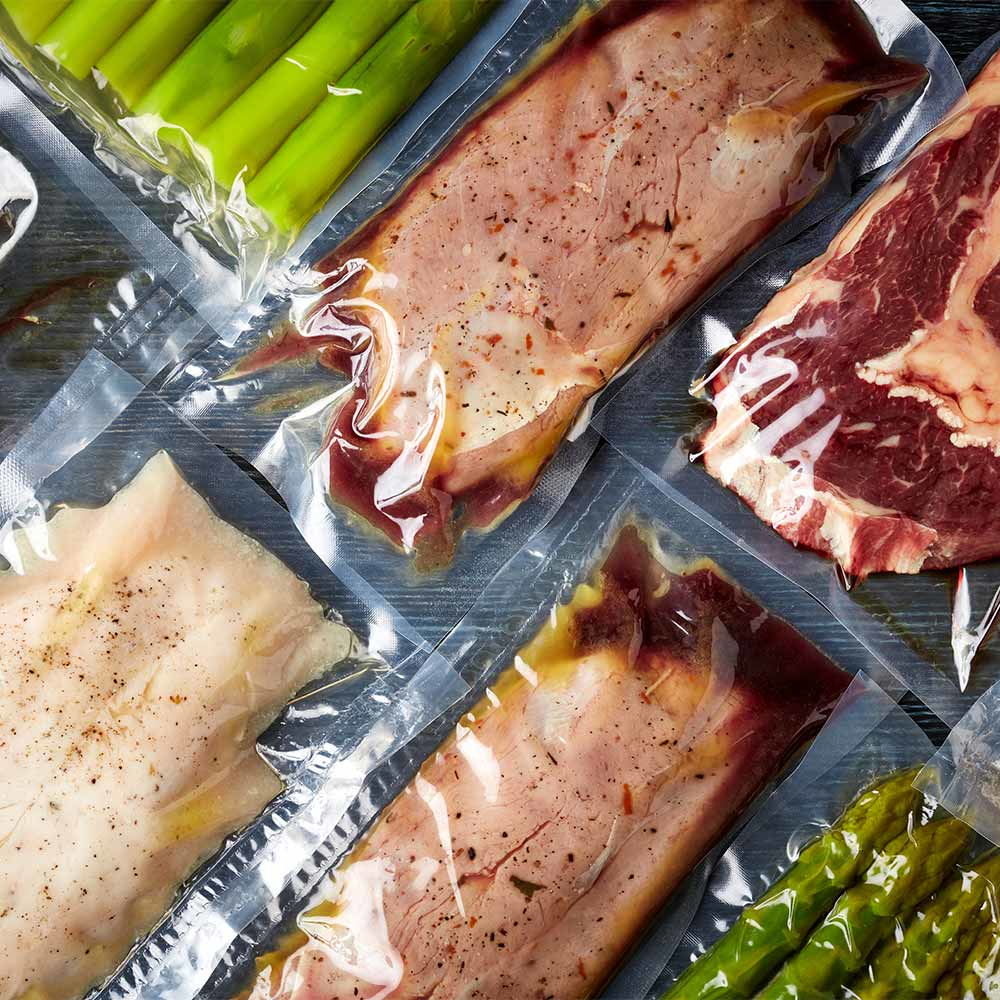 Vari pacchi sottovuoto di carne cruda e verdure su una superficie in legno.