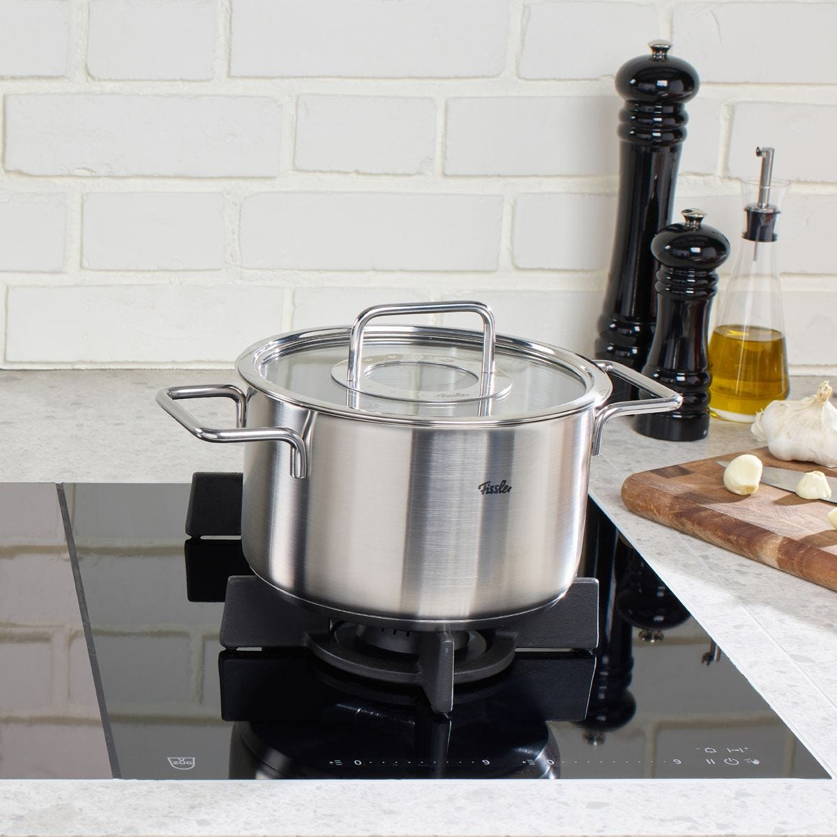Pentola in acciaio inox su fornello nero. Saliere e pepiere, olio, aglio e tagliere sullo sfondo.
