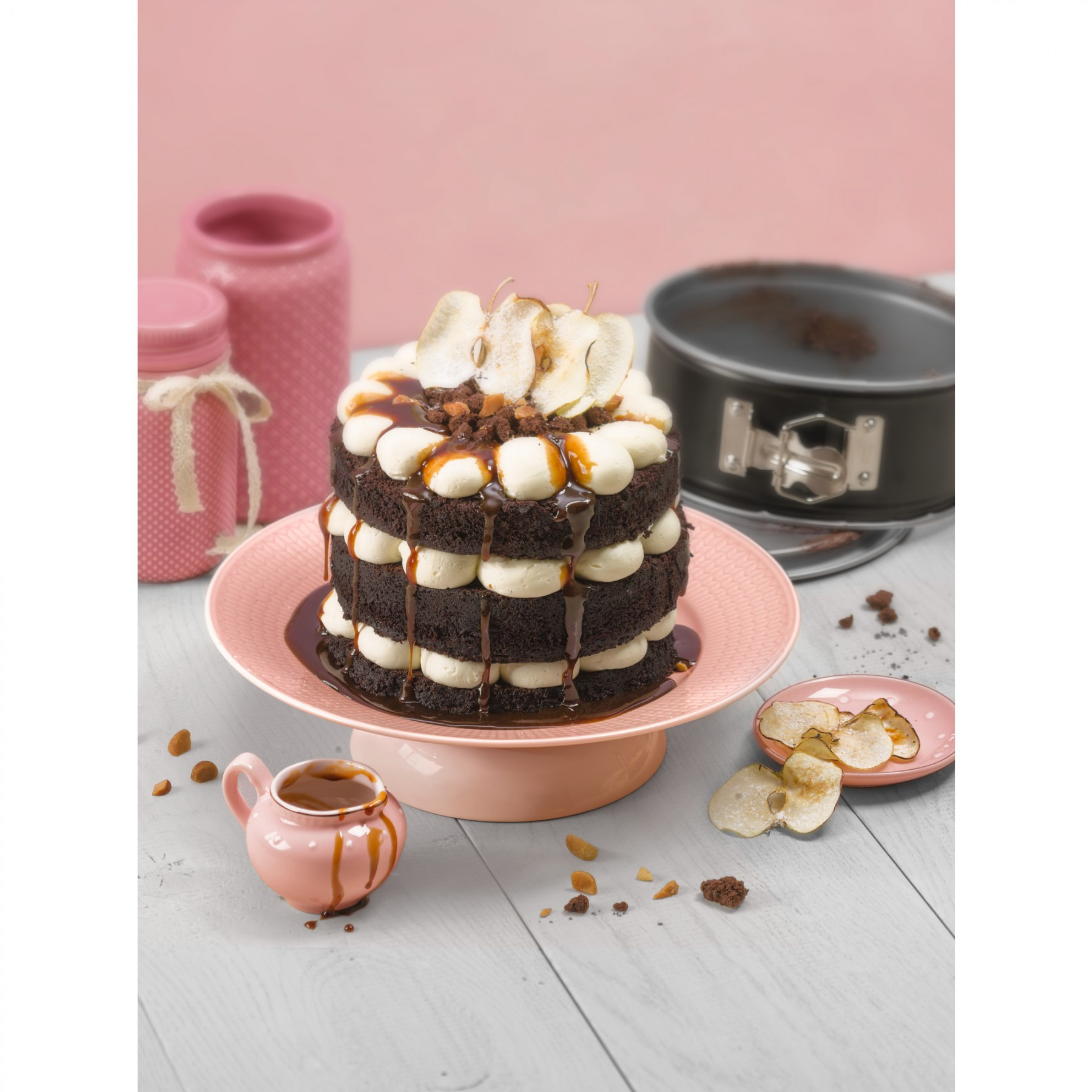 Warstwowe ciasto ze śmietaną, karmelem i plasterkami jabłka na różowym stojaku i tle.