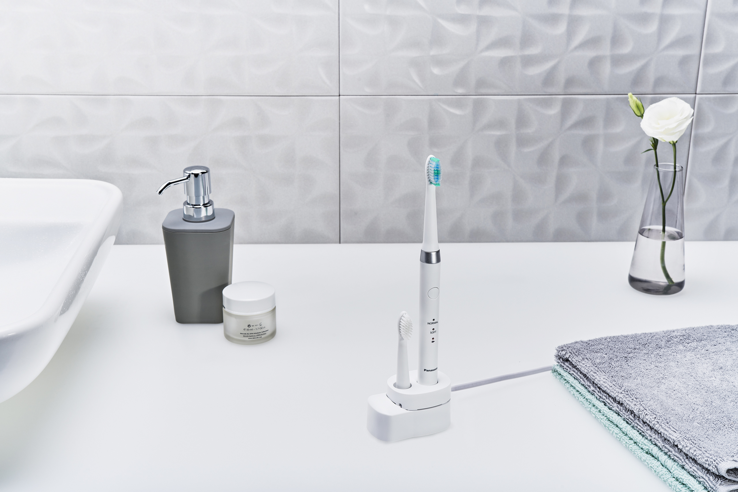 Blat łazienkowy ze szczoteczką do zębów, dozownikiem mydła, słoikiem kremu, kwiatem i ręcznikiem.