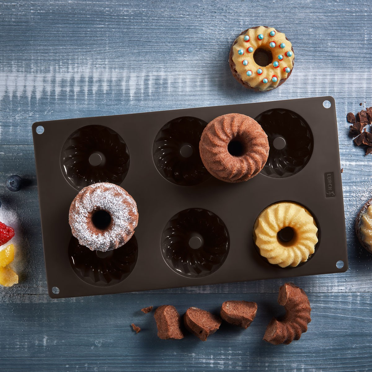 Donutförmige Kuchen in einer Silikonform auf einer blauen Holzoberfläche. Einige Kuchen sind mit Puderzucker bestäubt.