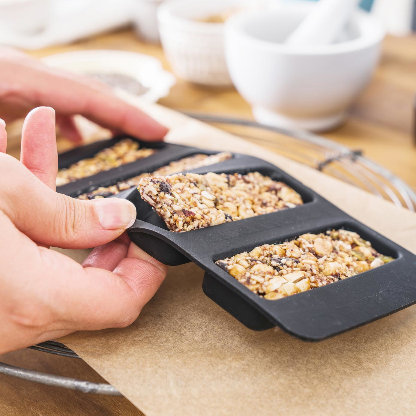 Mani che mettono barrette di granola in uno stampo rettangolare nero su carta marrone su una superficie di legno.