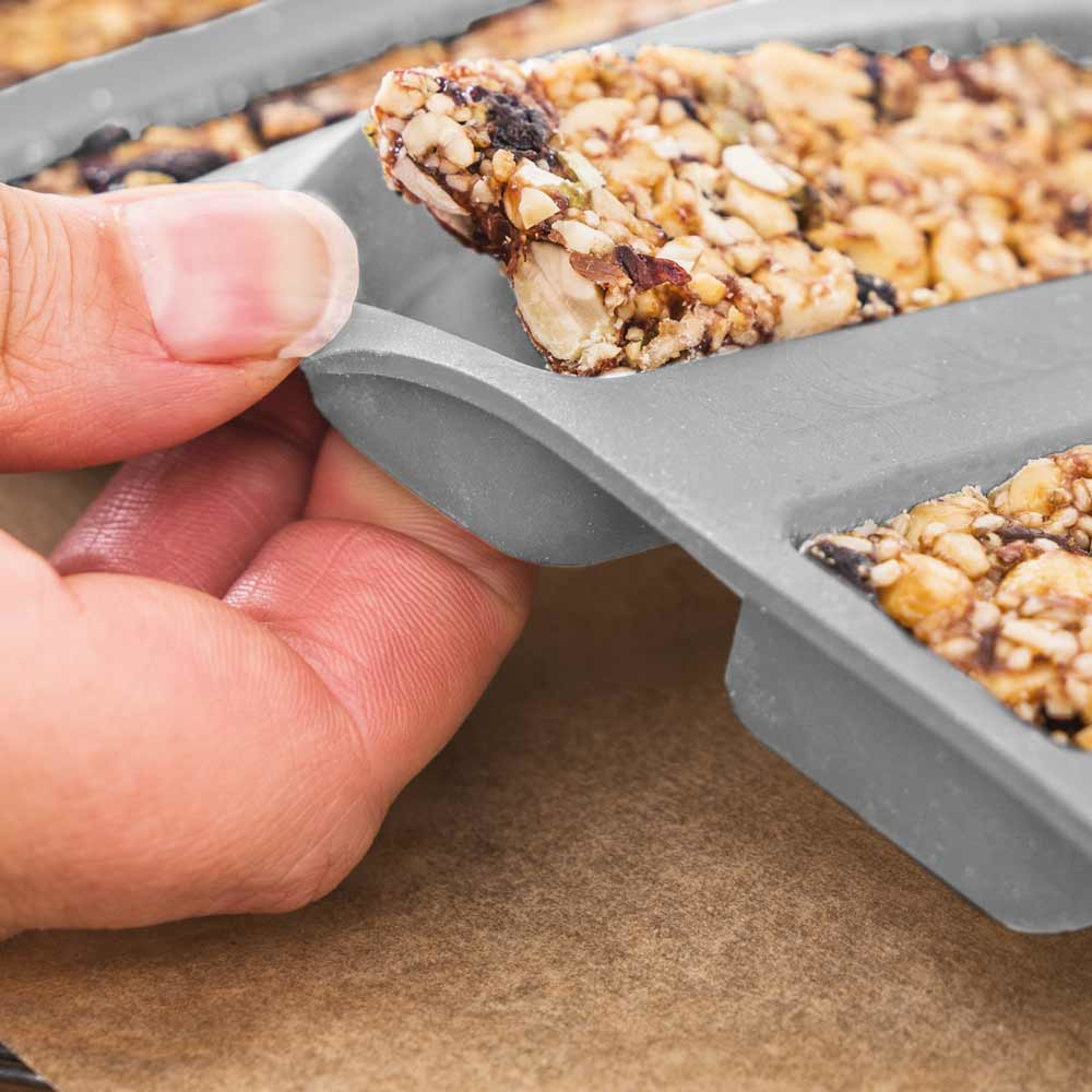 Una mano che rimuove un pezzo di barretta di granola da uno stampo grigio. La barretta di granola ha noci e semi. Lo sfondo è marrone.