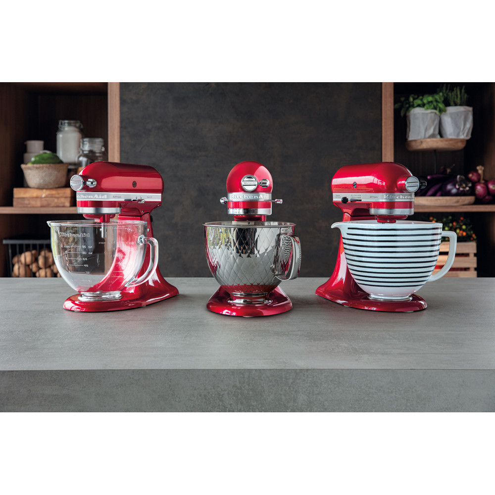 Gri bir tezgah üzerinde farklı kaseleri olan üç kırmızı KitchenAid mikseri. Arka plan rafları ve mutfak eşyalarını içerir.