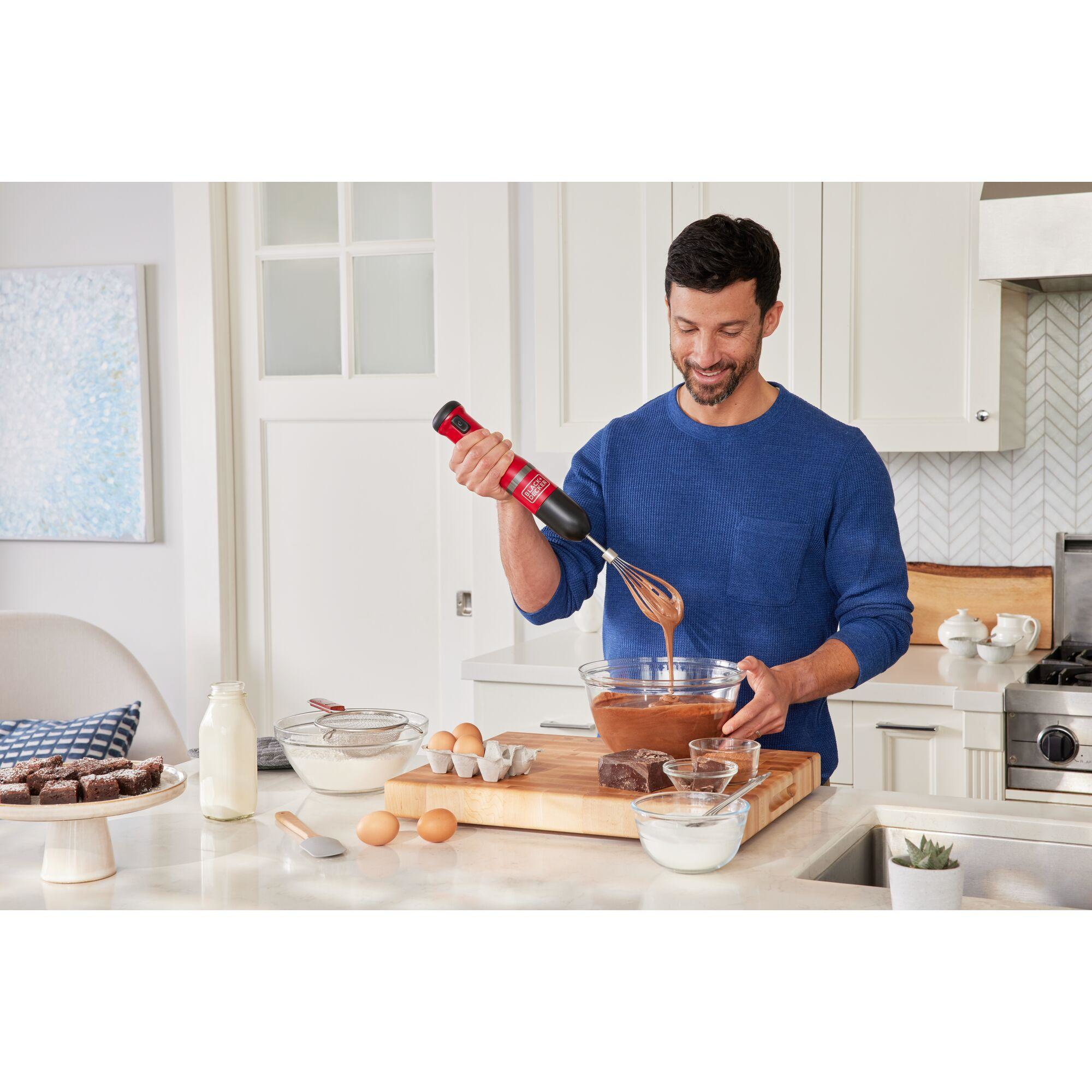 Un hombre mezcla masa con una batidora de mano roja en una cocina, ingredientes en la encimera.