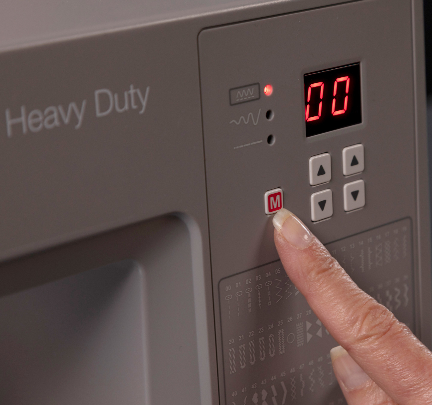 Maszyna do szycia z palcem naciskającym przycisk 'M'. Wyświetlacz pokazuje '00'.