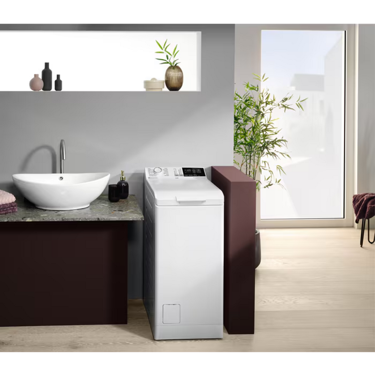 Biała pralka w łazience, z umywalką, roślinami i oknem.