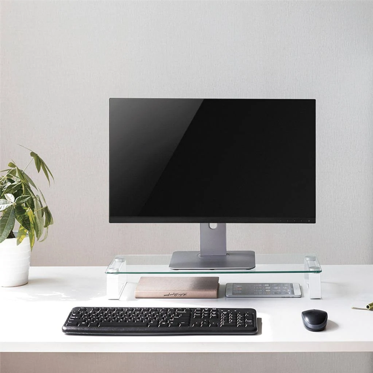 Konfiguracja komputera z monitorem, klawiaturą i szklaną podstawką pod monitor na białym biurku.