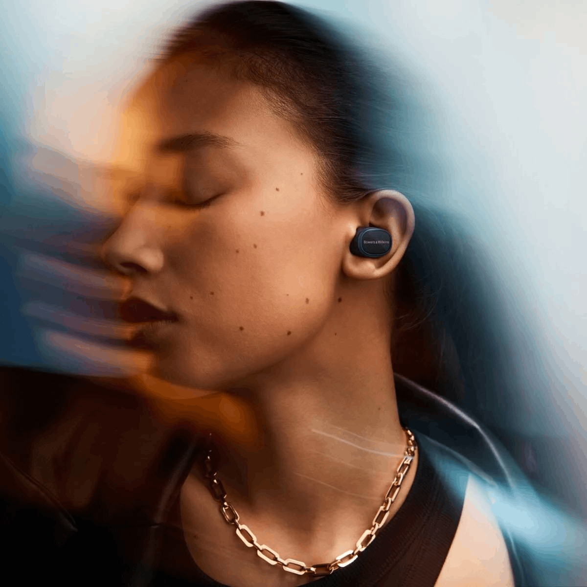 Profil einer Frau, die Bowers & Wilkins Ohrhörer und eine goldene Kette trägt, mit abstrakten Lichteffekten.