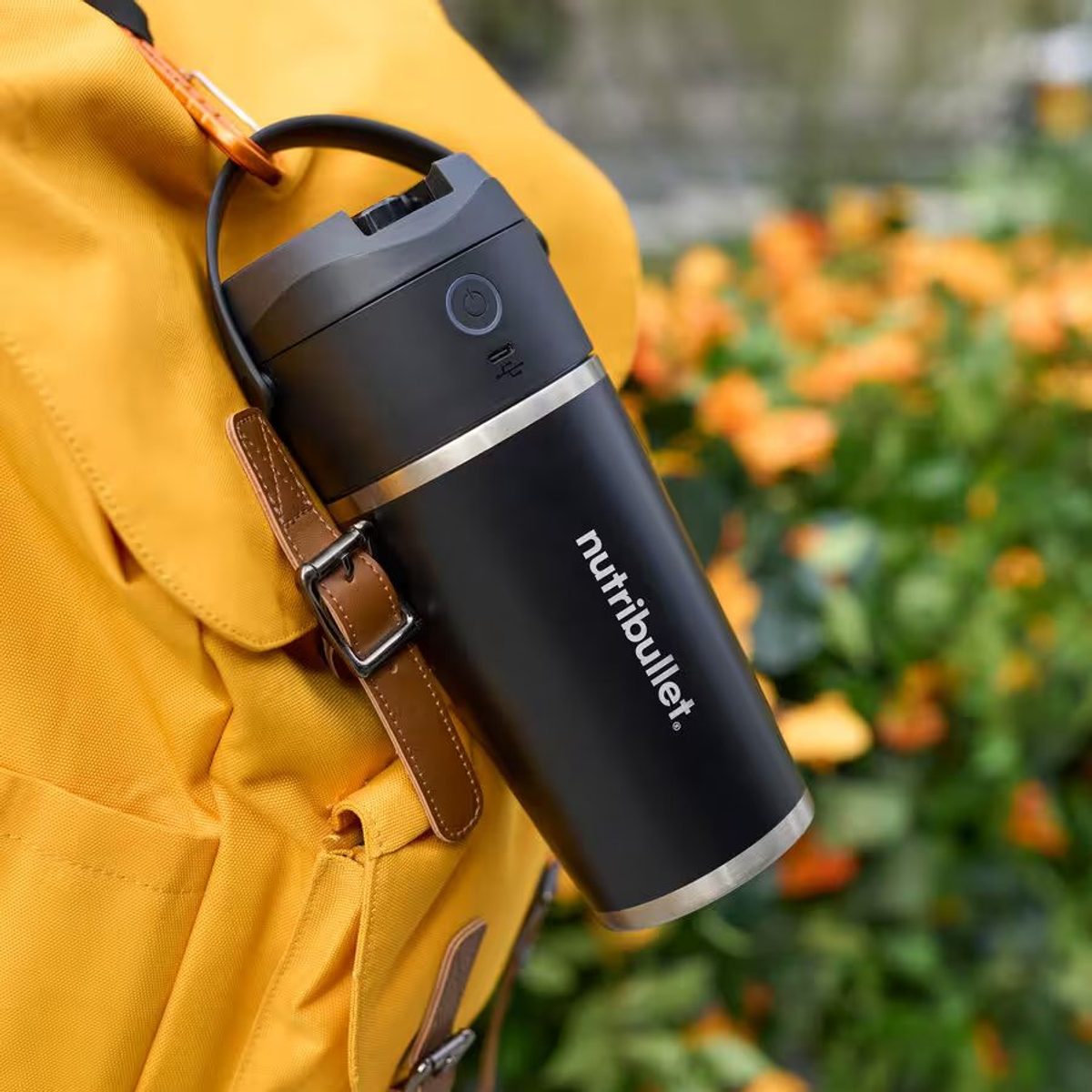 Schwarze und silberne Wasserflasche an einem gelben Rucksack mit orangefarbenen Blumen im Hintergrund.