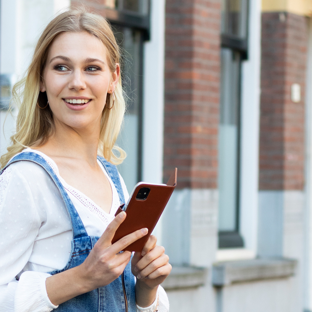 Blonde Frau, die eine braune Handyhülle hält, lächelt, mit einem Gebäude im Hintergrund.