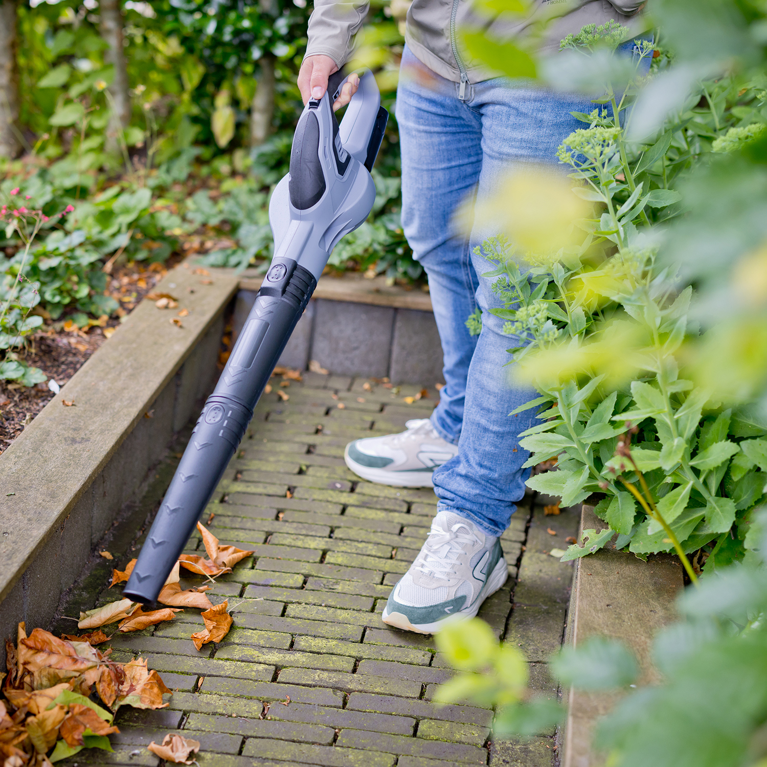 Persoon die een grijze bladblazer gebruikt op een bakstenen pad, omringd door groene planten.