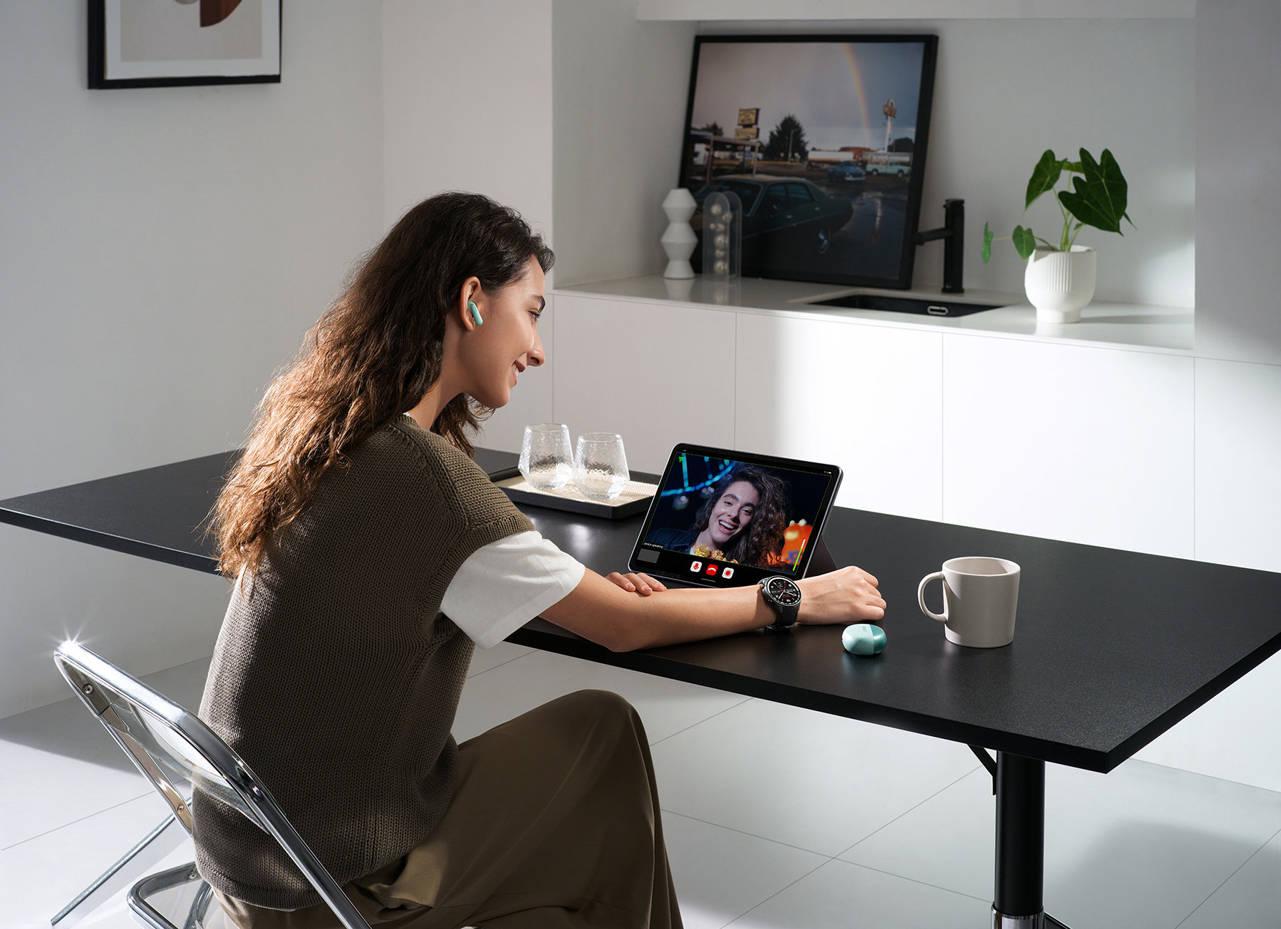 Kobieta z słuchawkami używa tabletu do wideo rozmowy. Kubek i zielone słuchawki na stole.