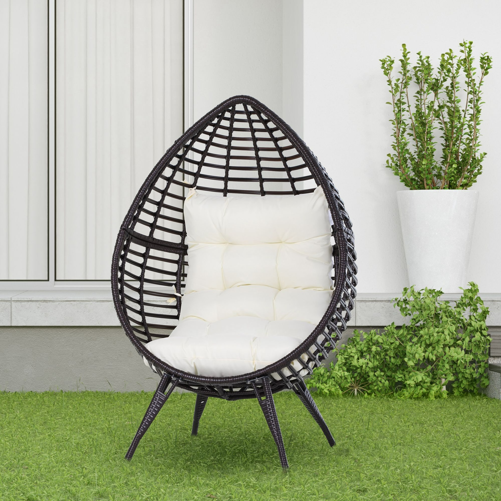 Weiden-Eiersessel mit Kissen auf Gras, Pflanzen im Hintergrund, weiße Wand.