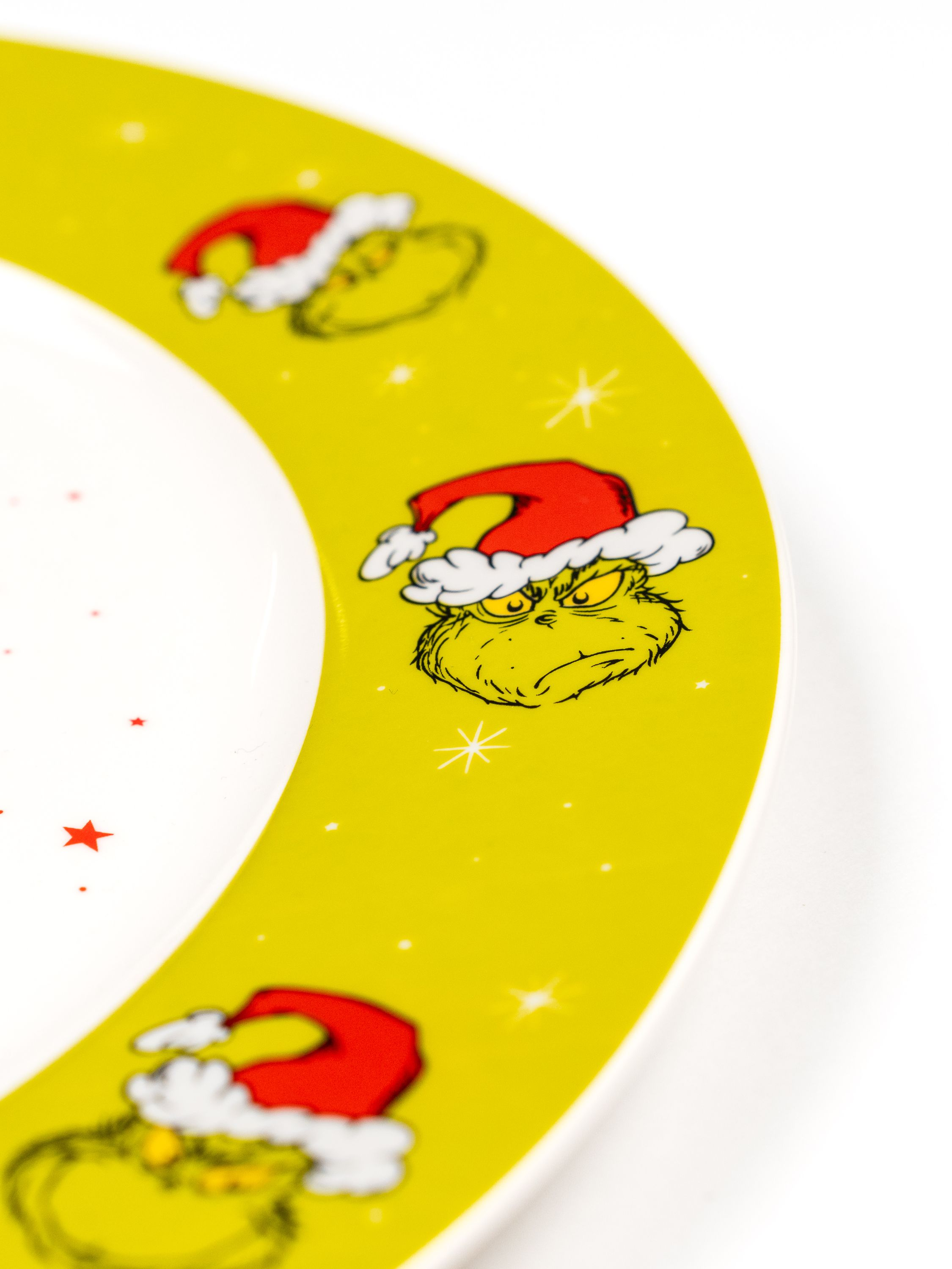 Teller mit Grinch-Gesichtern mit Weihnachtsmützen. Gelber Rand, weißer Mittelteil. Rote Sterne im Hintergrund.