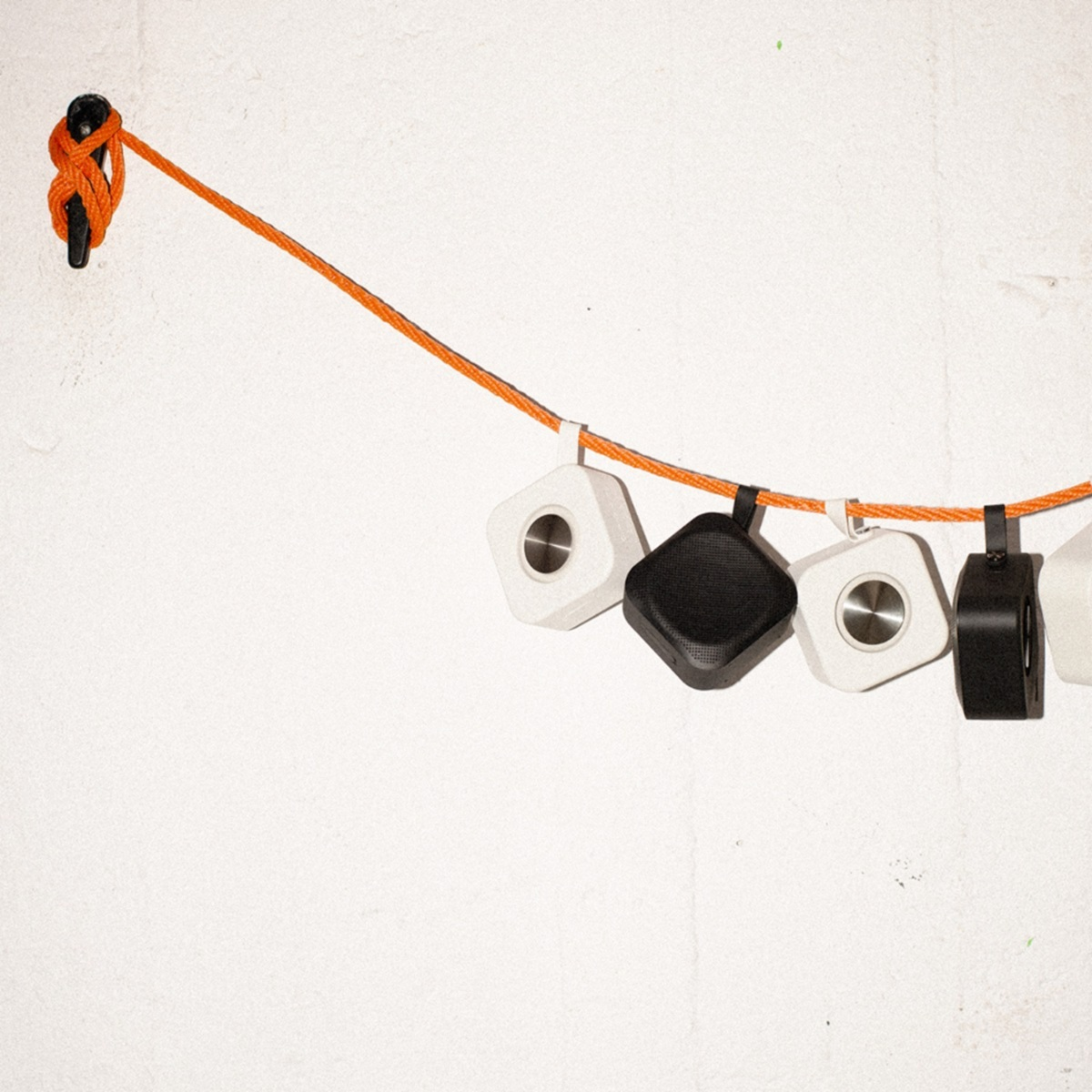 Głośniki wiszące na pomarańczowym sznurze na białej ścianie. Głośniki są białe, czarne i szare.