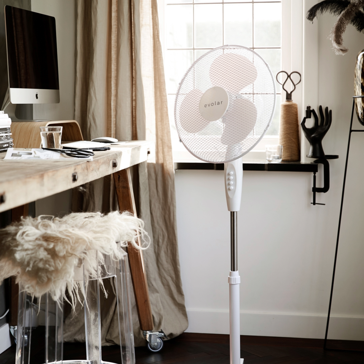 A standing fan in a room with a desk, computer, and window. Neutral tones.