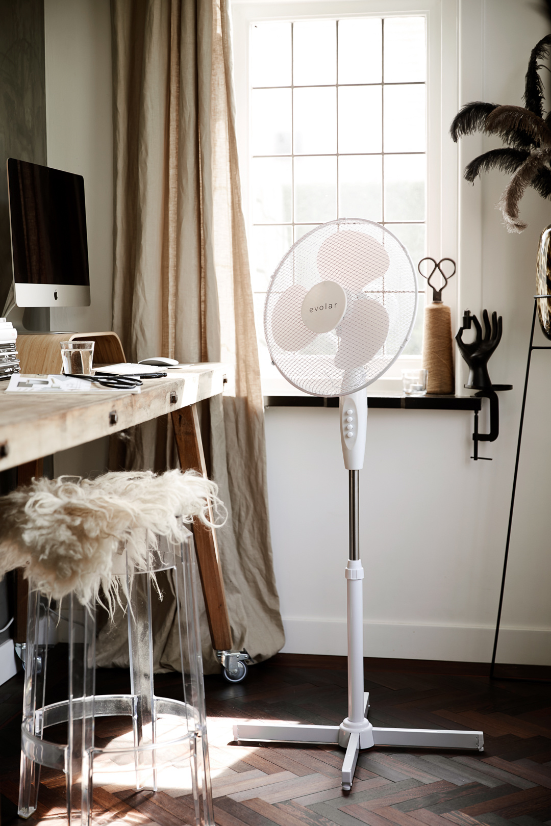 A white standing fan in a room with a wooden desk, stool, and window.