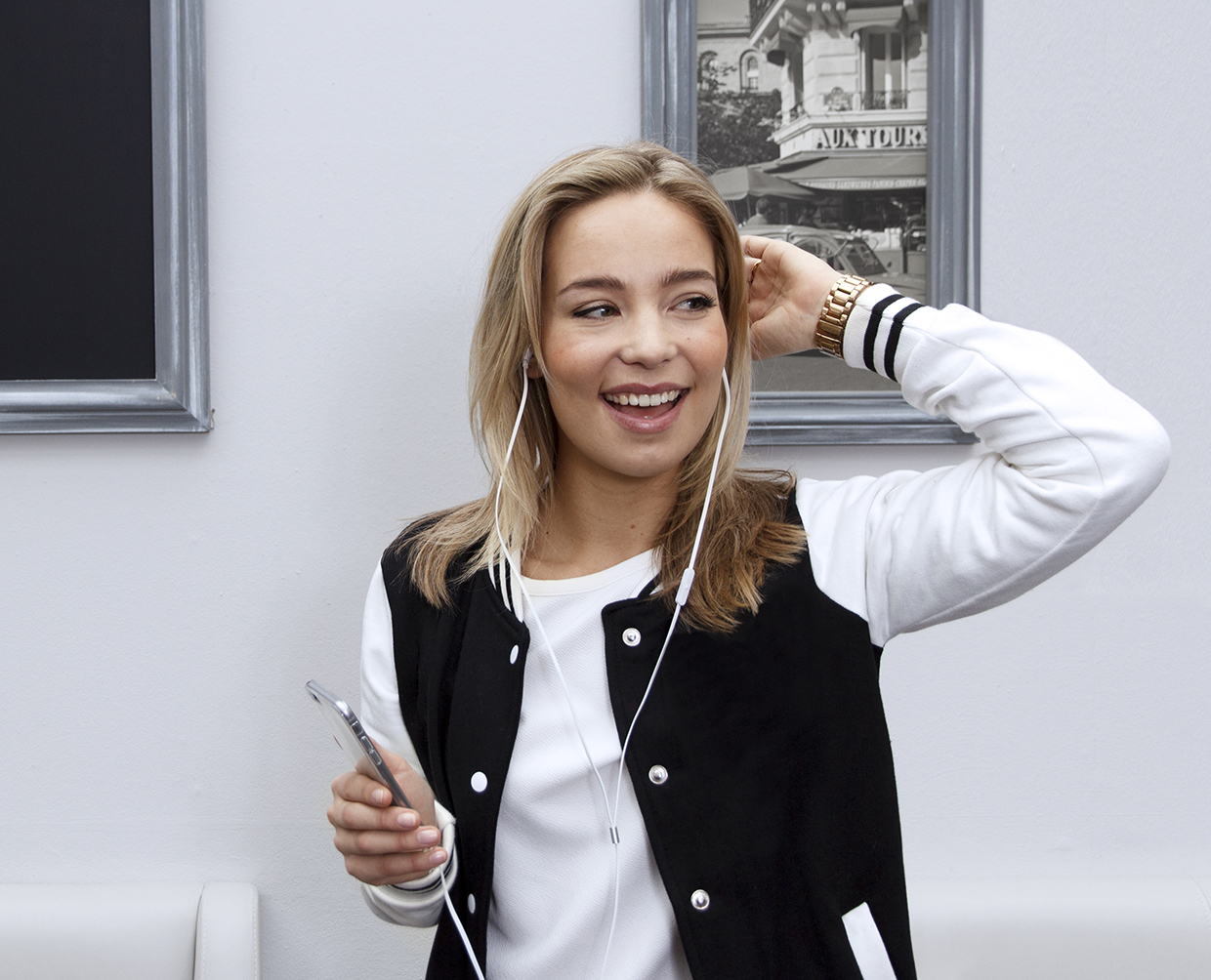 Blonde vrouw lacht, houdt een telefoon vast, draagt oortelefoons en een jas, op een witte muur.