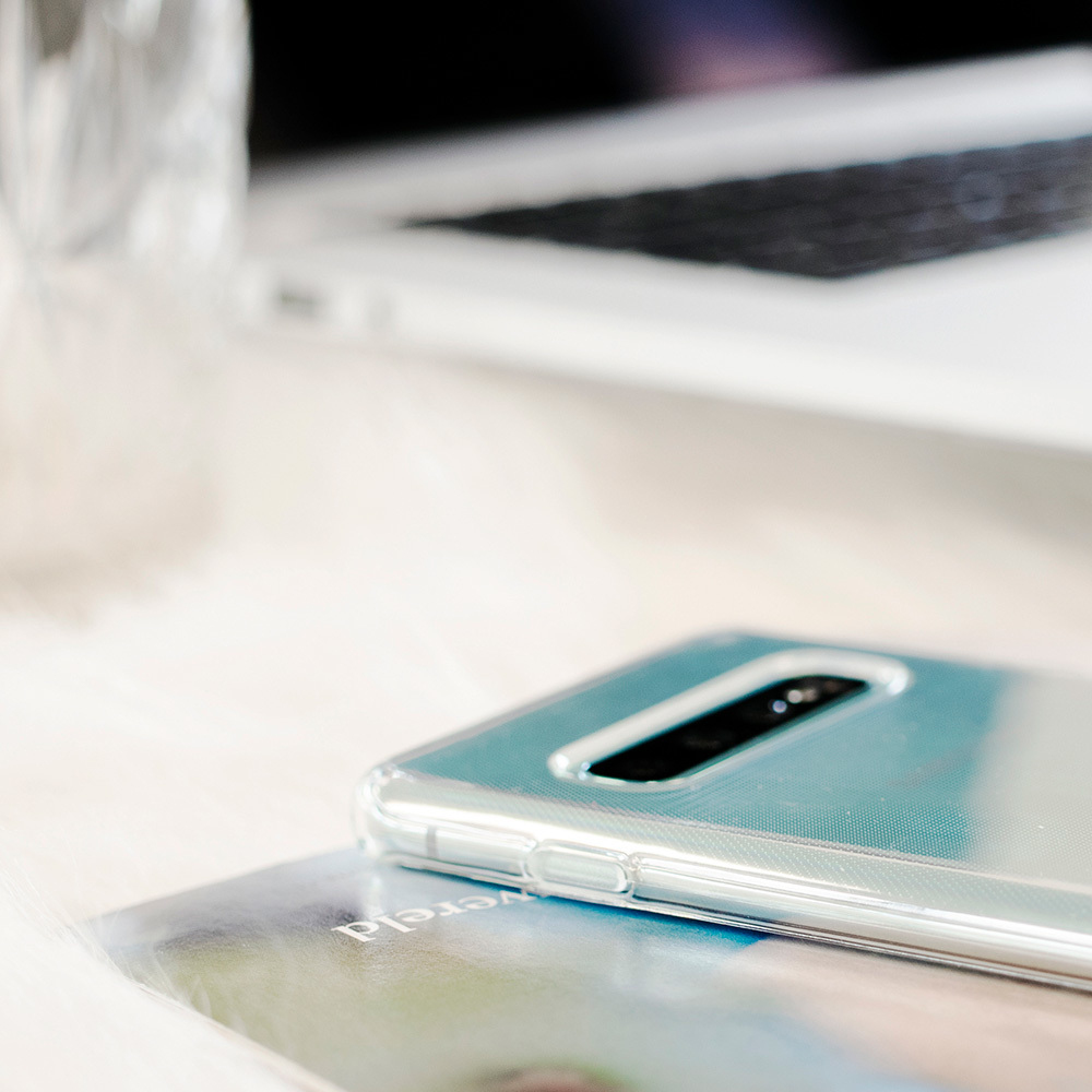 Een telefoon in een doorzichtige hoes op een tijdschrift, met een laptop en glas op de achtergrond, een heldere omgeving.