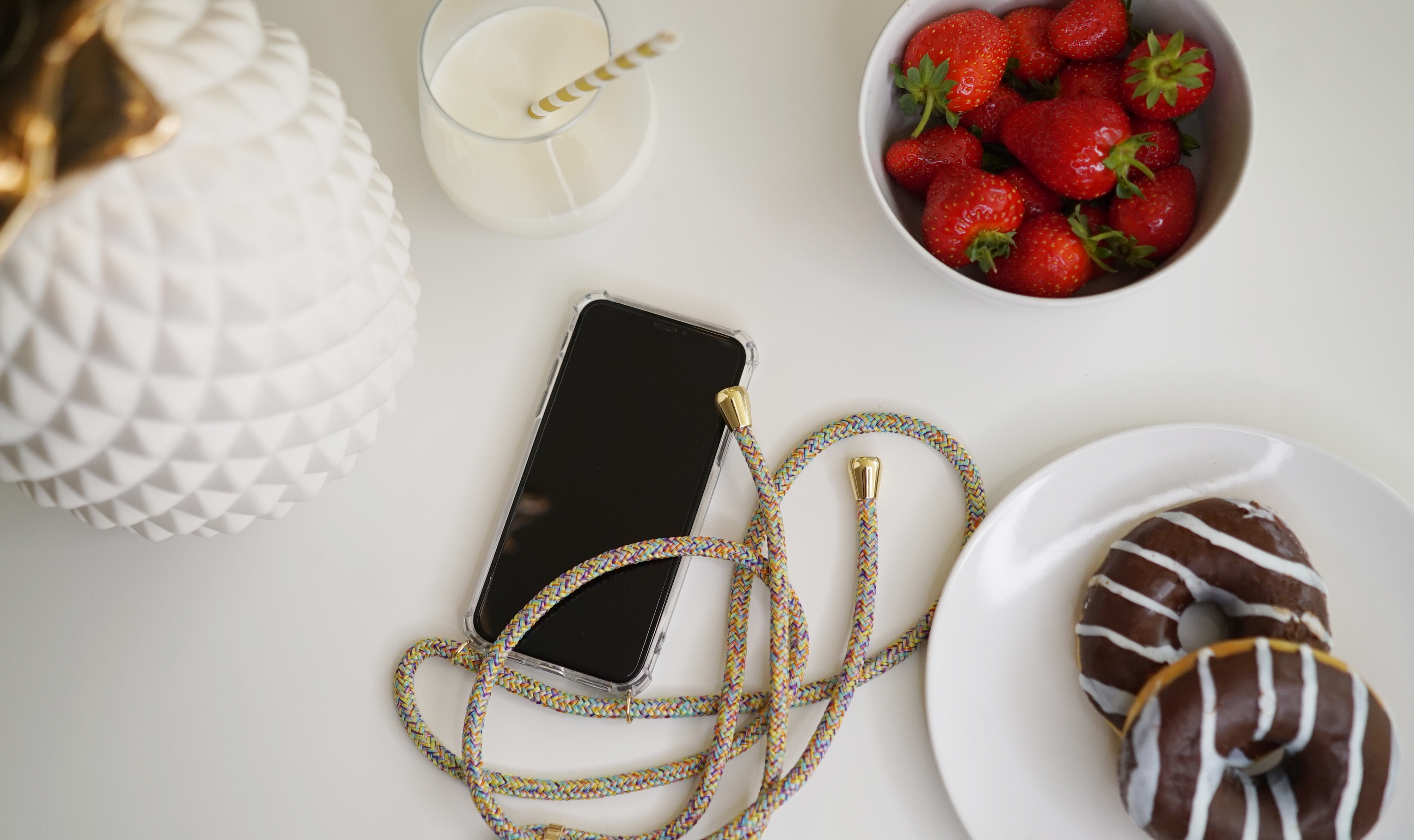 Draufsicht, Telefon mit Lanyard, Glas Milch, Erdbeeren und Donuts auf weißem Tisch.