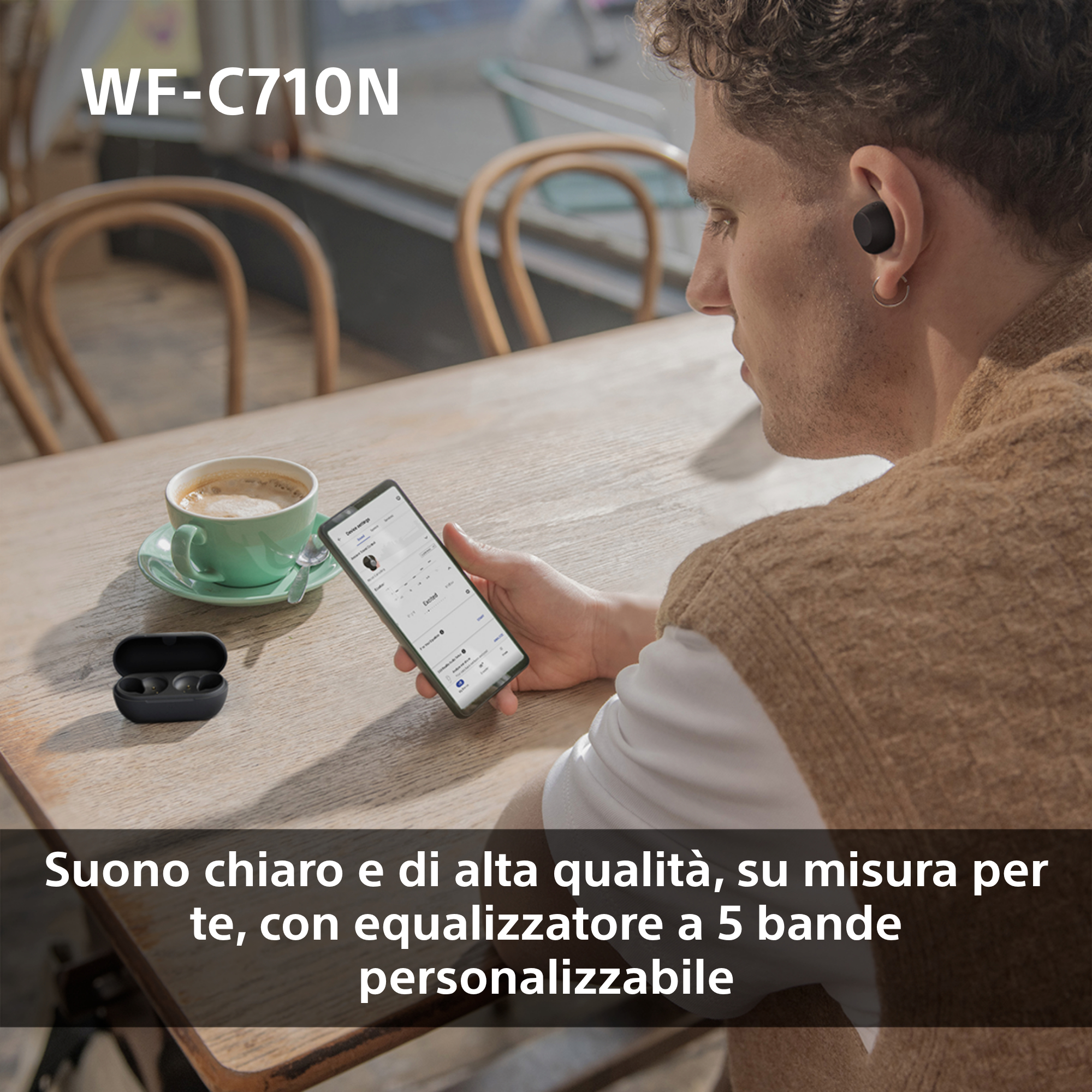 Un uomo usa auricolari mentre guarda uno smartphone, con una tazza di caffè e custodia accanto.