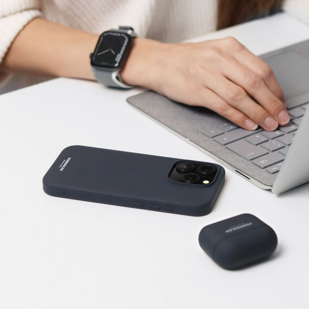 Die Hand einer Person auf einem Laptop. Ein Telefon, eine Uhr und eine Kopfhörerhülle liegen auf einem weißen Tisch.