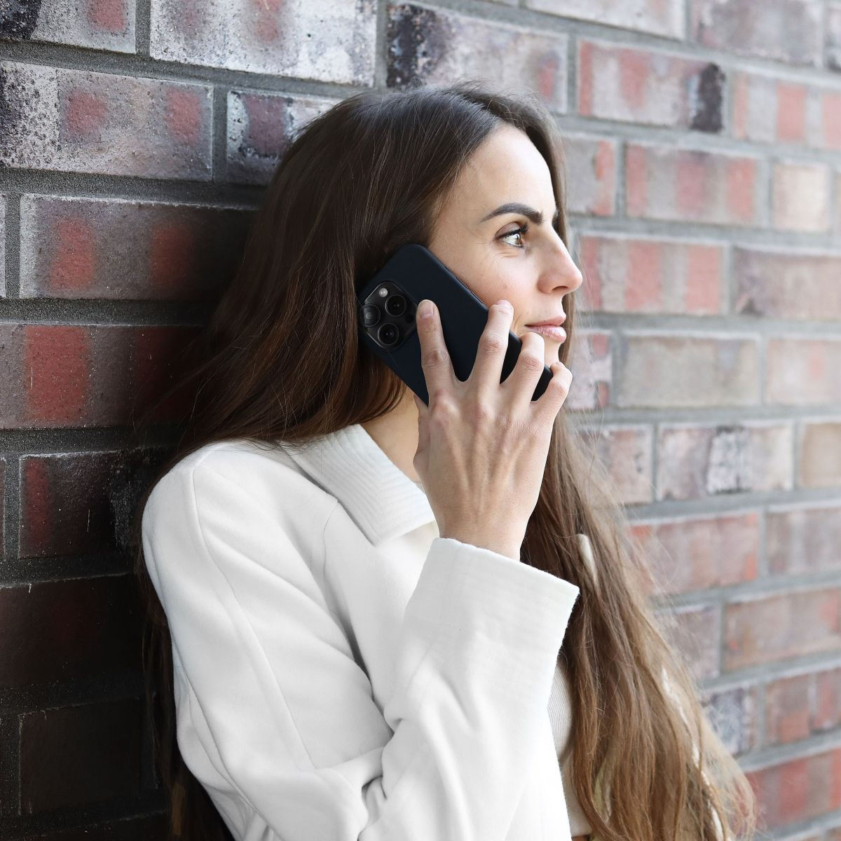 Frau telefoniert. Sie lehnt sich an eine Backsteinmauer und trägt eine weiße Jacke.