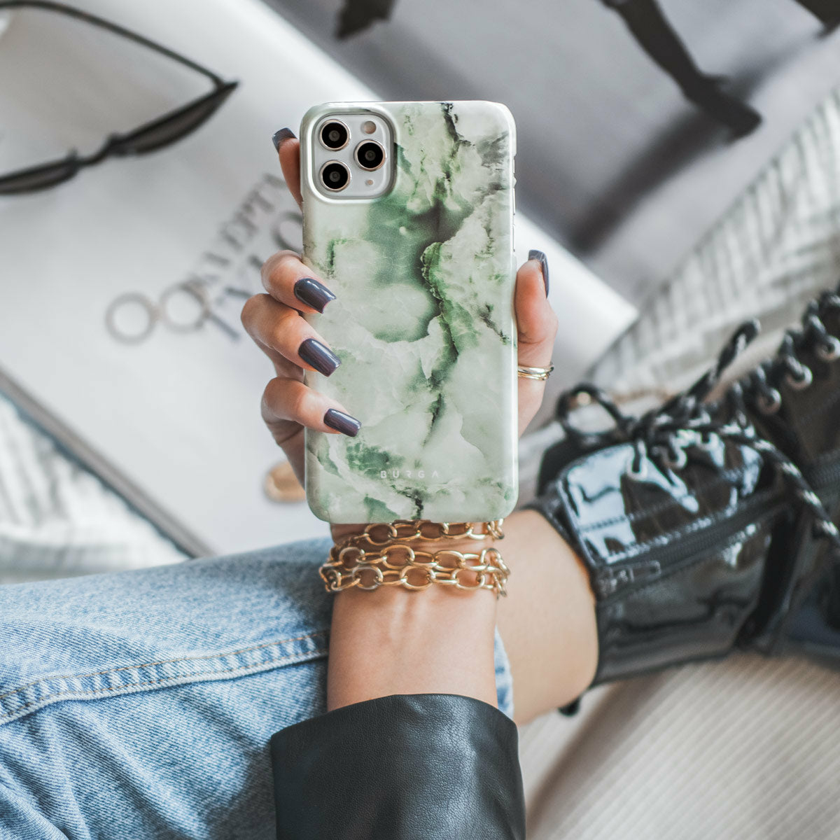 Una persona tiene in mano una custodia per telefono verde marmorizzata, con braccialetti d'oro e jeans.